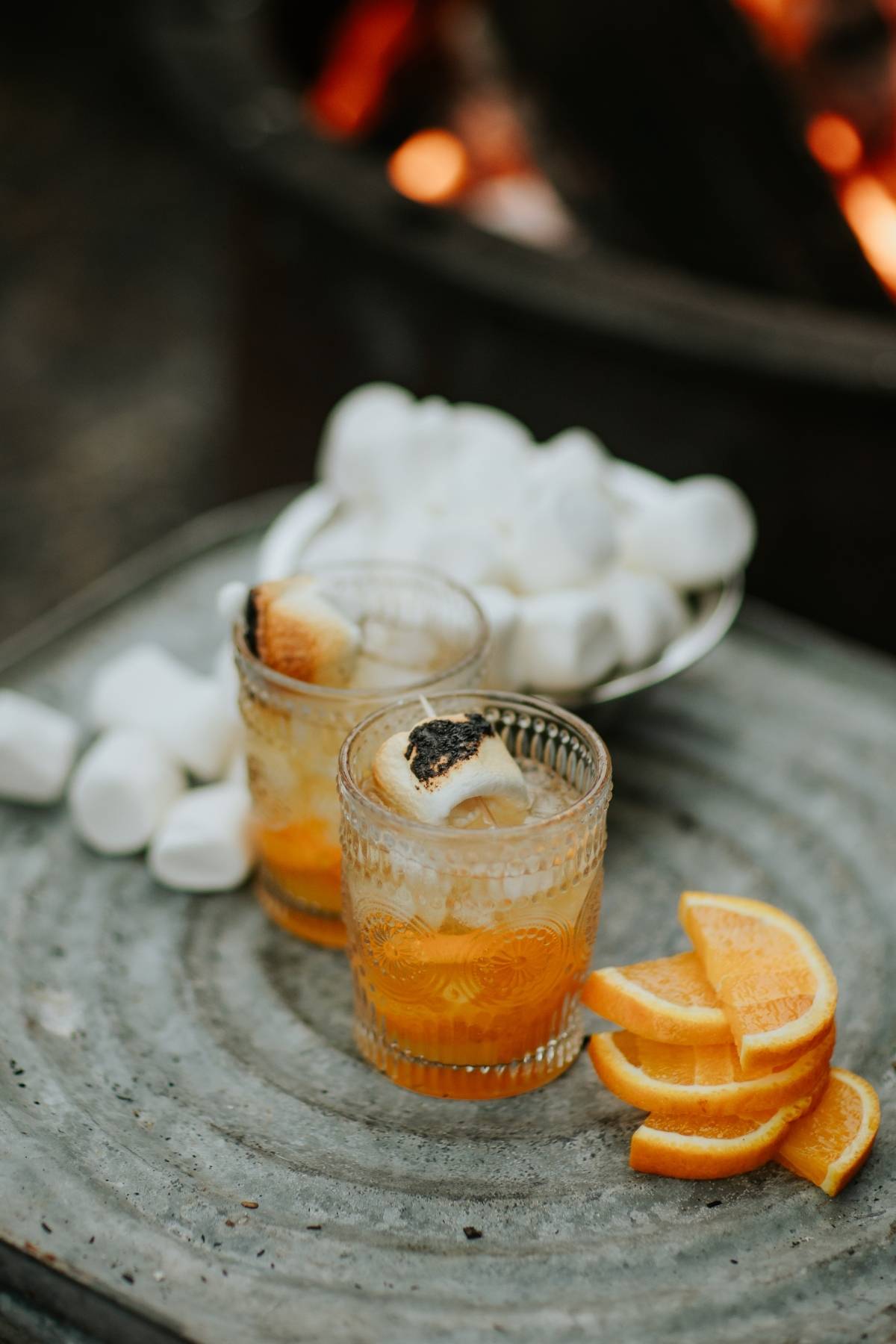 Two glasses with toasted marshmallows, orange slices, and marshmallows on a tray near a fire.