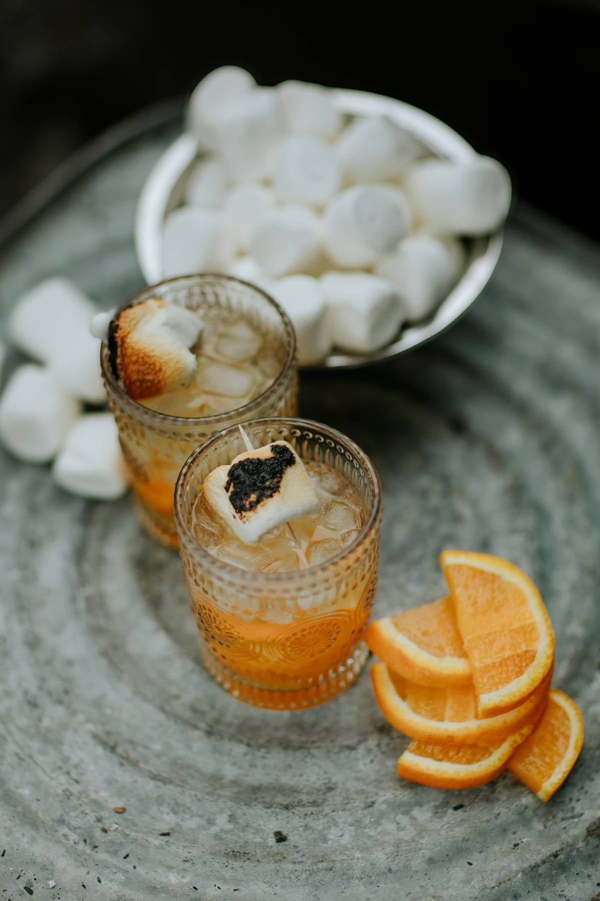 Two glasses with orange drinks topped with toasted marshmallows, orange slices, and a bowl of marshmallows nearby.