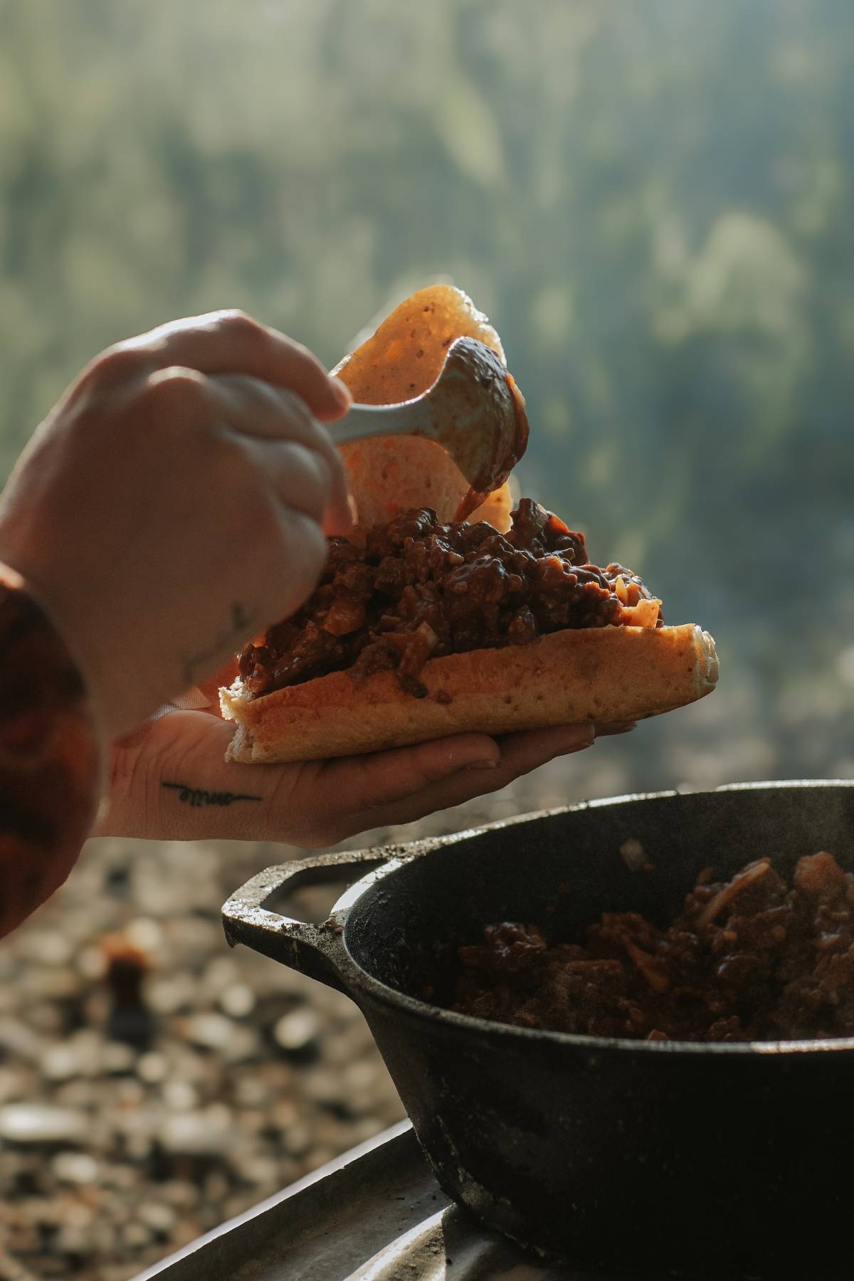 A person spoons stew onto a sandwich bun outdoors next to a cast iron pan.