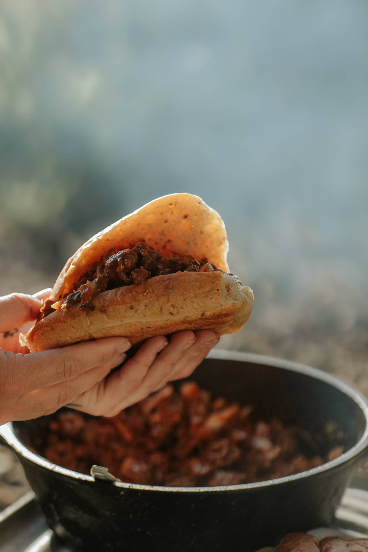 A hand holds a taco shell filled with a chunky, saucy filling above a pot outdoors.