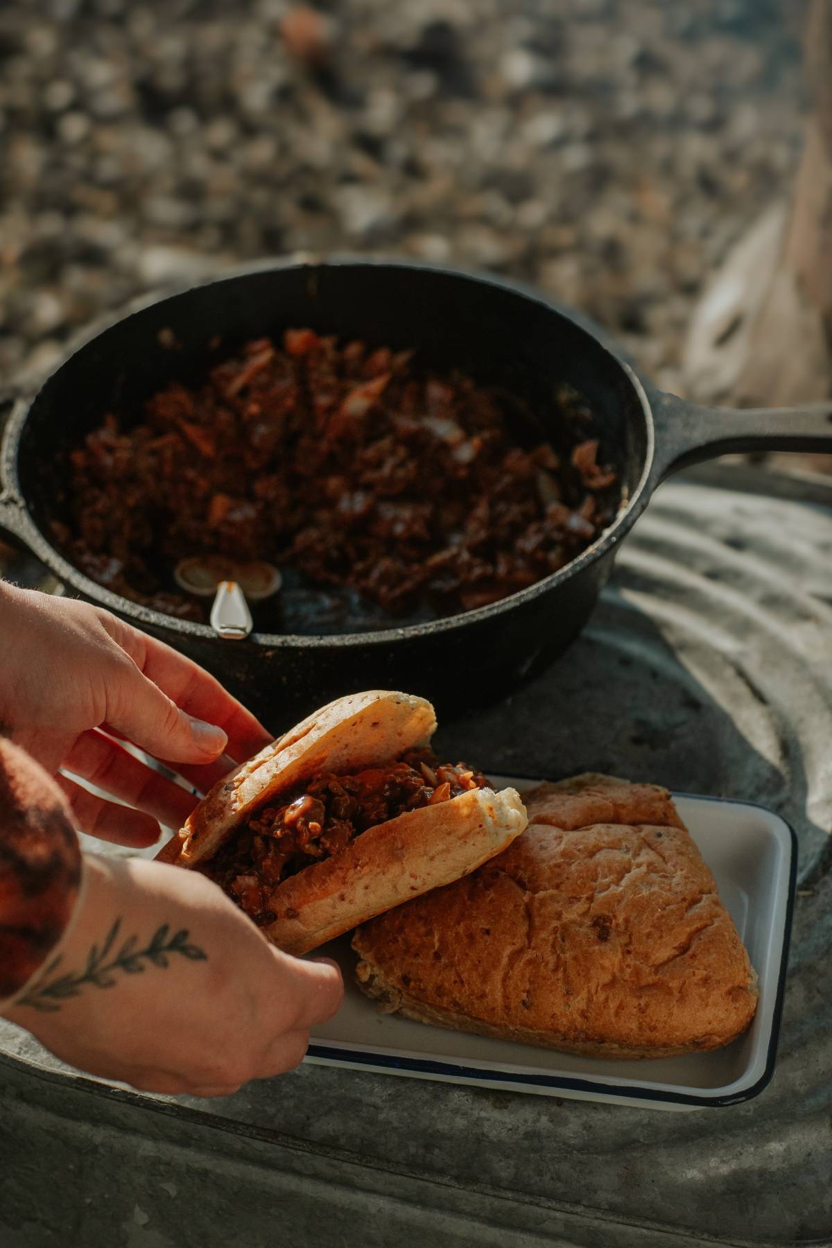 Hands assemble a sandwich filled with stew beside a pan of cooked meat on an outdoor surface.