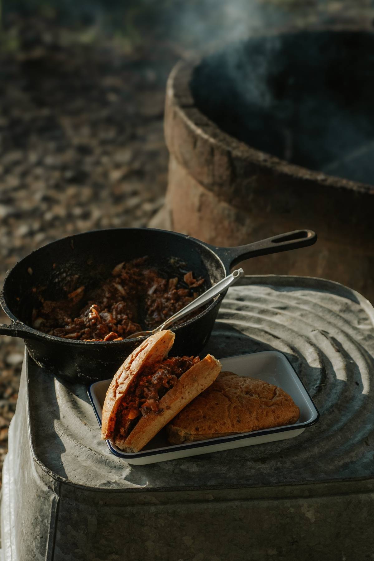 Cooked meat in a cast iron skillet and a sandwich with meat filling on a tray near a fire pit outdoors.