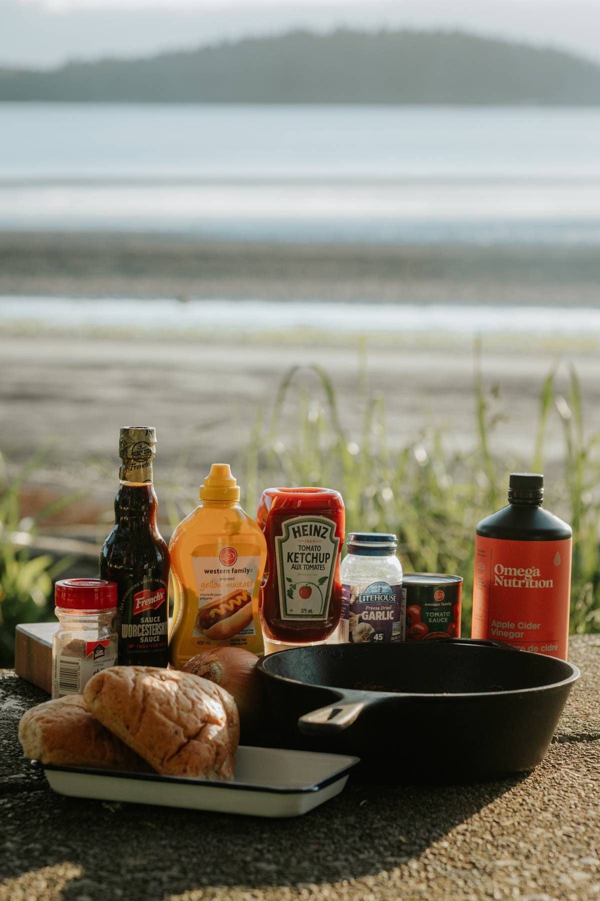 Burger ingredients and condiments arranged outdoors on a table with a beach and water in the background.