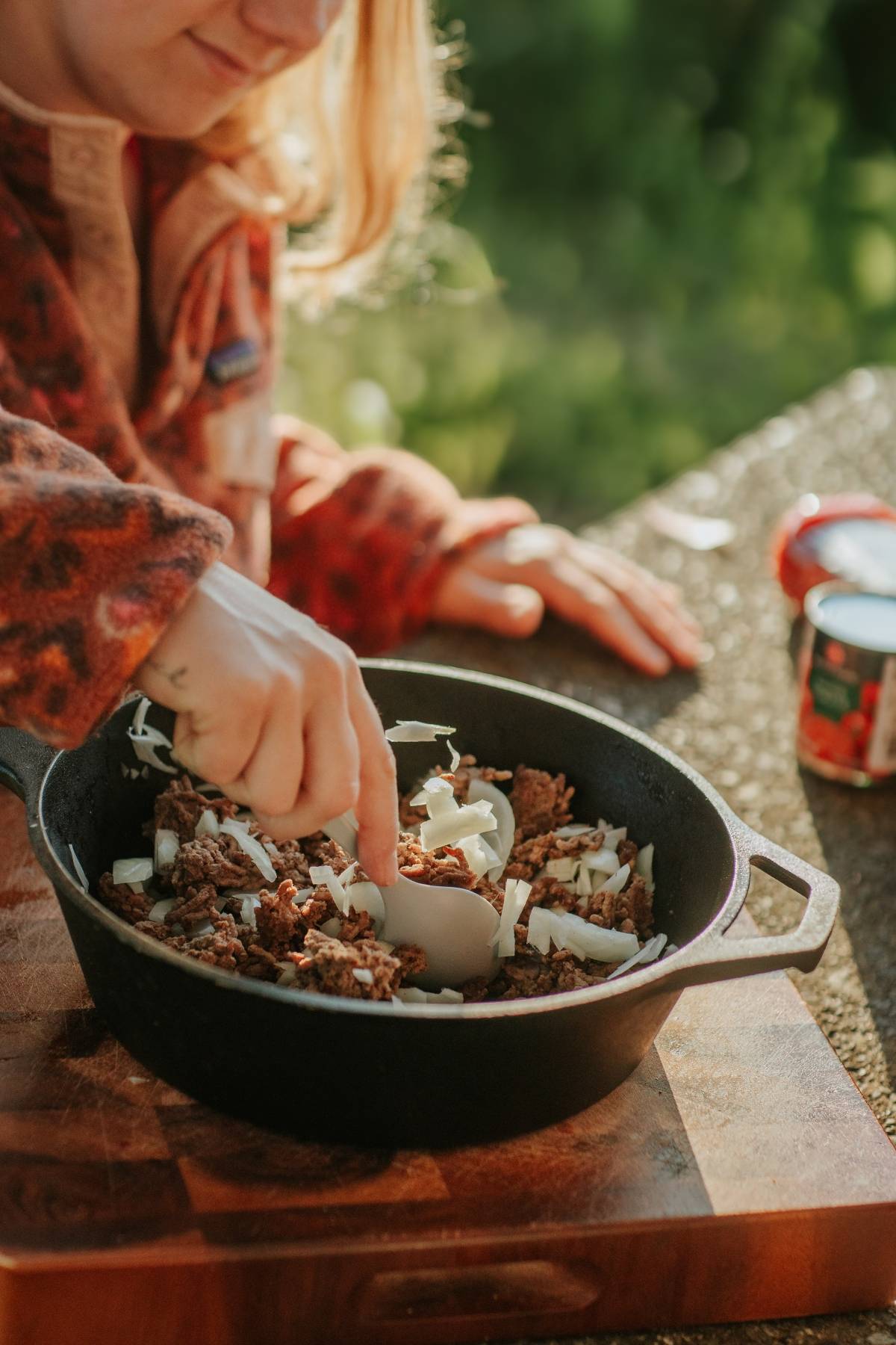 Person adding chopped onions to ground meat in a black skillet outdoors on a wooden surface.