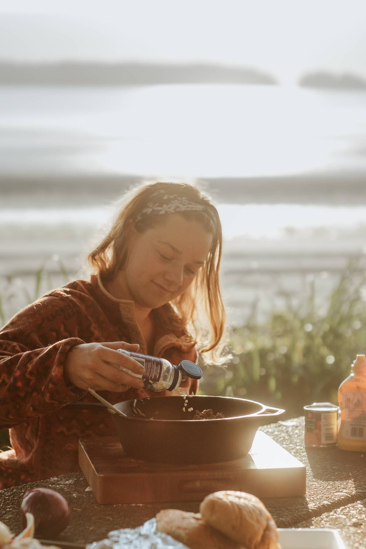 Woman seasoning food in a pan at an outdoor table, with a beach and ocean in the background at sunset.