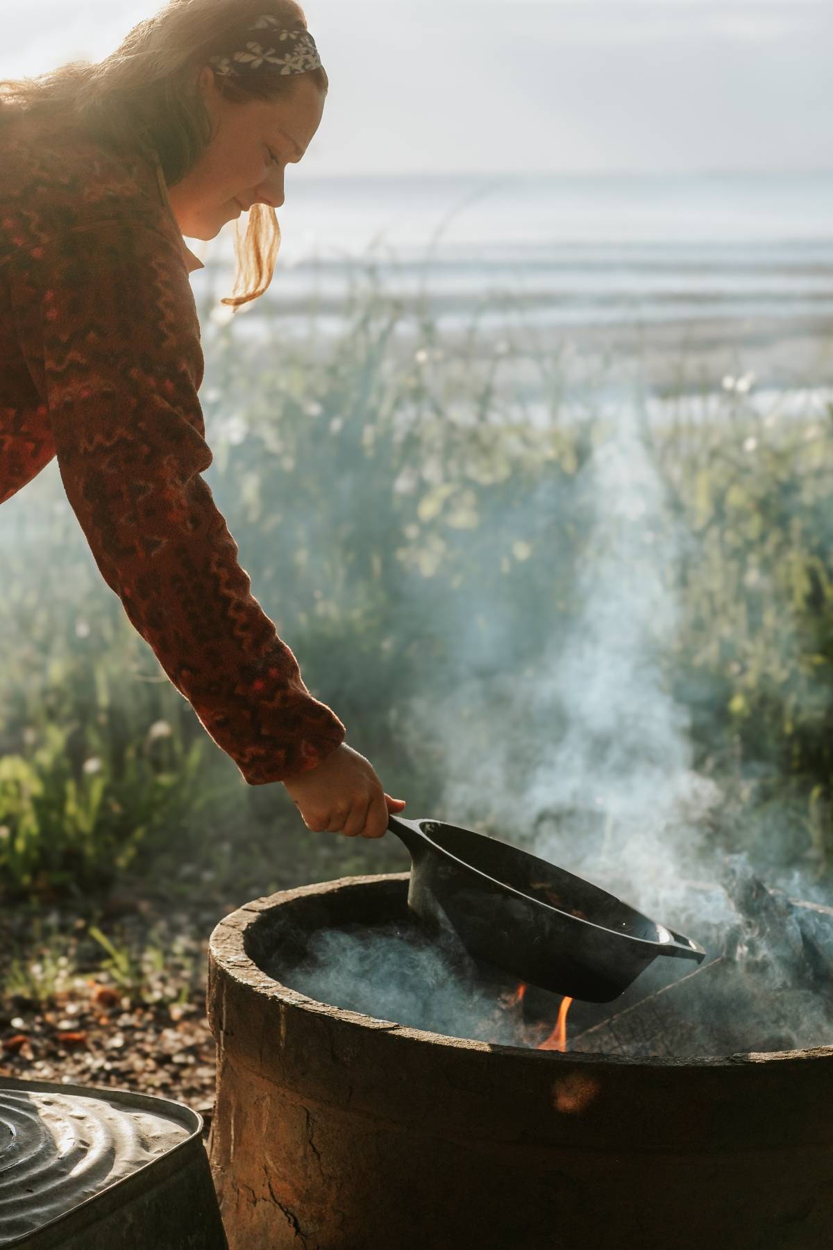 Woman cooking with a cast iron skillet over an open fire outdoors near a lake.
