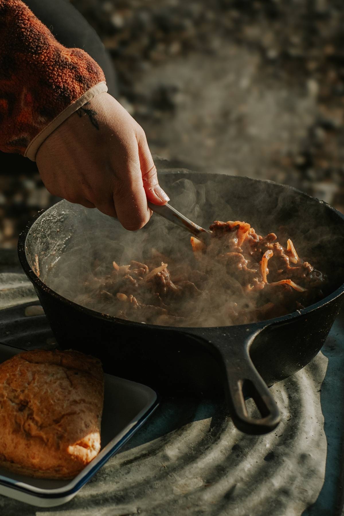 A hand stirs steaming food in a cast iron pan, with a biscuit on a plate nearby.