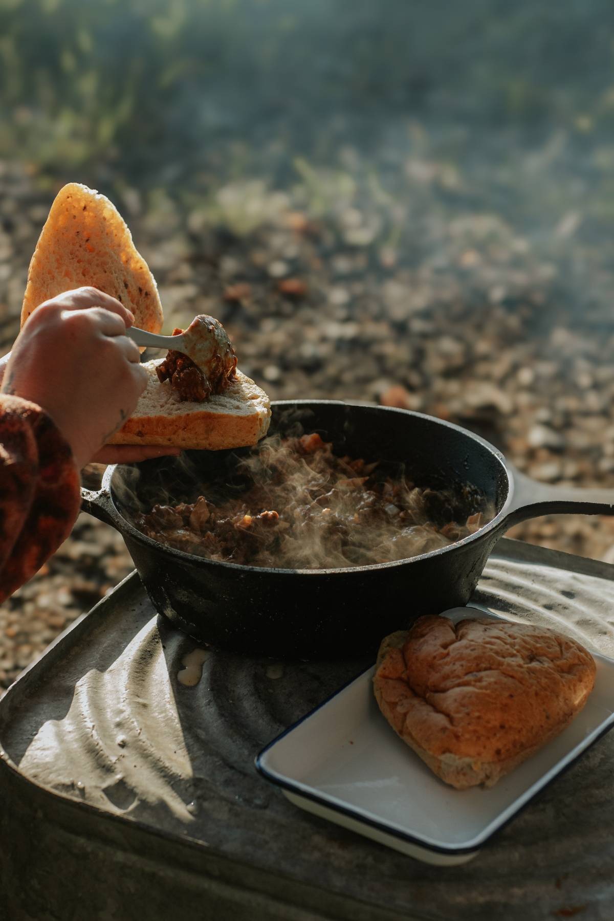 A person scoops hot stew from a cast iron pan onto bread outdoors, with another bread on a tray nearby.