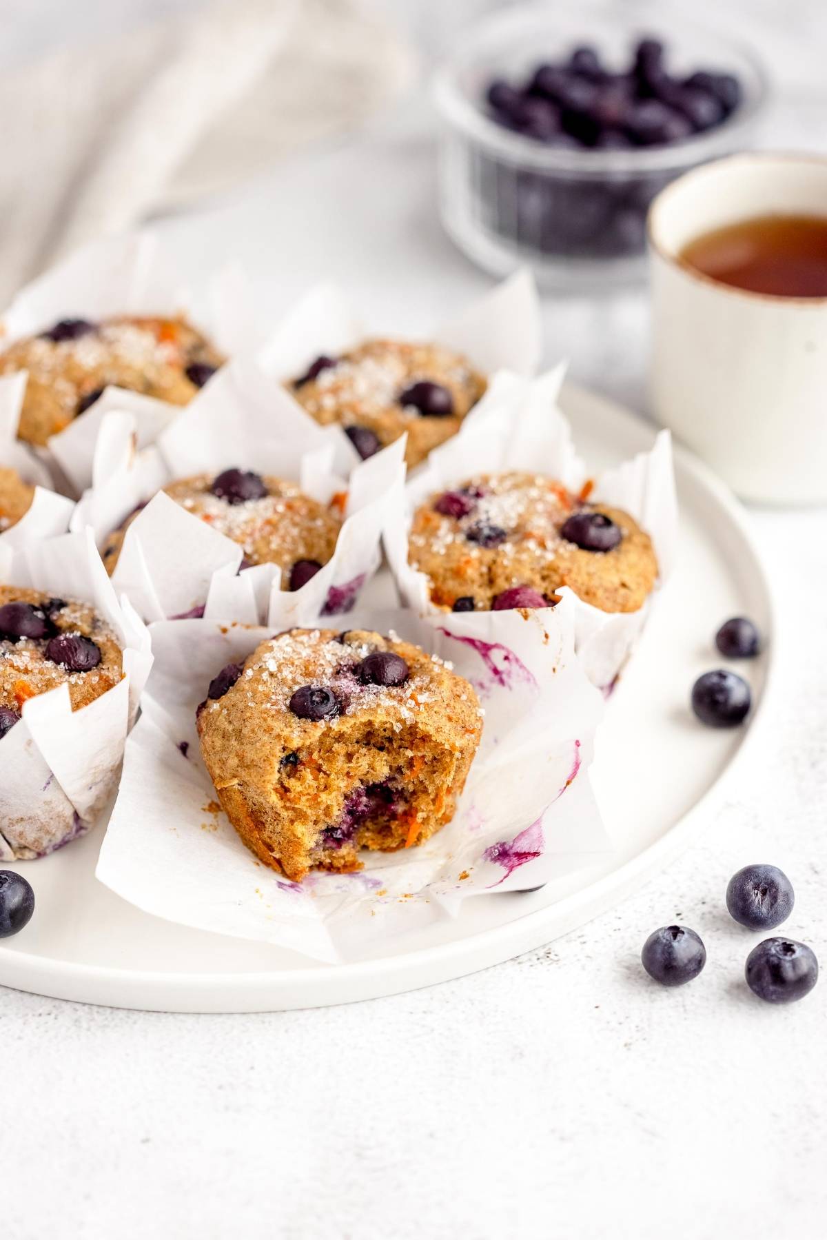 Healthy blueberry muffin with carrots served on a plate in parchment tulip muffin liners, accompanied by a cup of tea and fresh blueberries in the background.