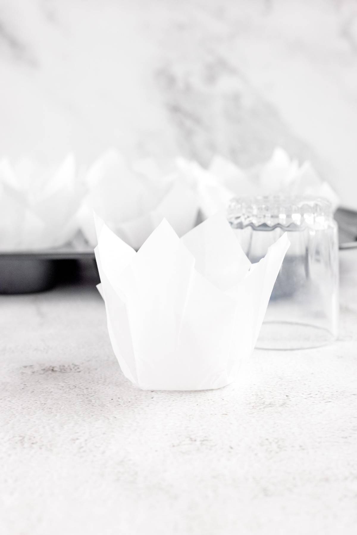 A parchment tulip muffin liner sits beside an upside-down glass and a muffin tin on a light surface, ready for your favorite carrot blueberry muffins.