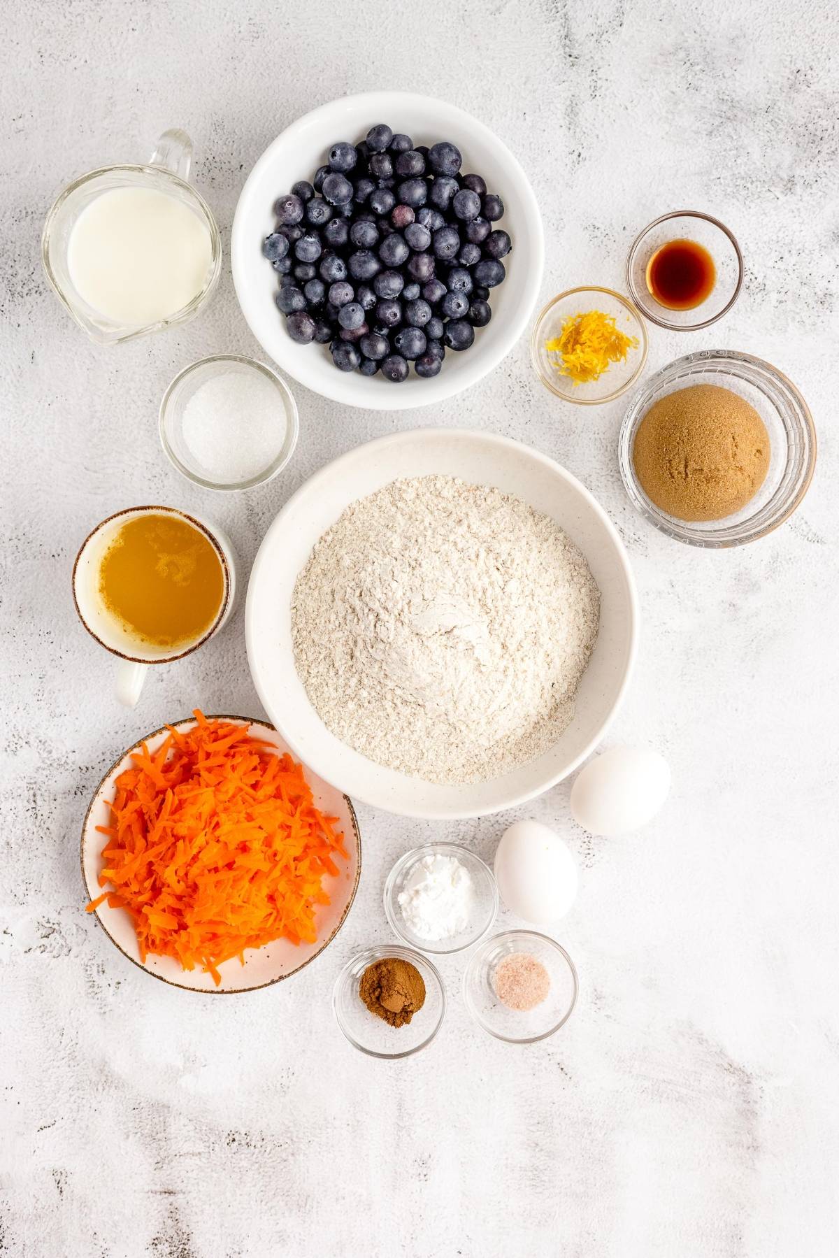 Baking ingredients on a white surface, including blueberries, flour, shredded carrots, eggs, and spices—perfect for a delicious carrot and blueberry recipe or healthy blueberry muffin with carrots.