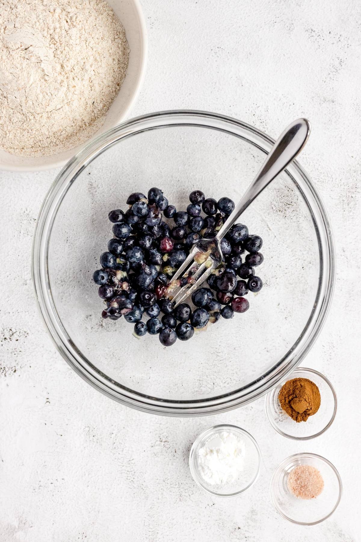 Clear glass bowl with blueberries and a fork, surrounded by flour, cinnamon, salt, and starch in small bowls—perfect for starting your favorite carrot blueberry muffins or another carrot and blueberry recipe.