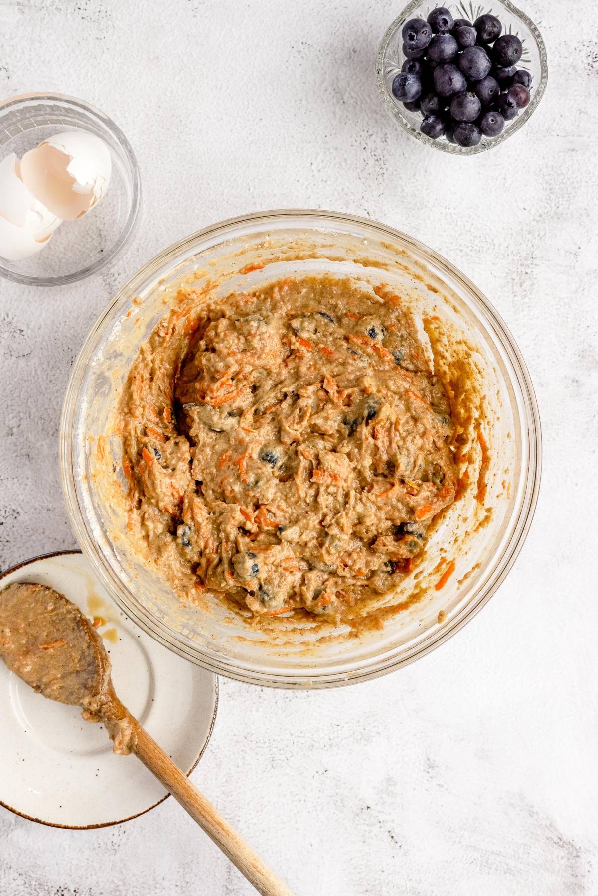A bowl of healthy blueberry muffin with carrots batter, featuring shredded carrots and blueberries, sits beside eggshells and a bowl of blueberries—perfect for your next carrot and blueberry recipe.