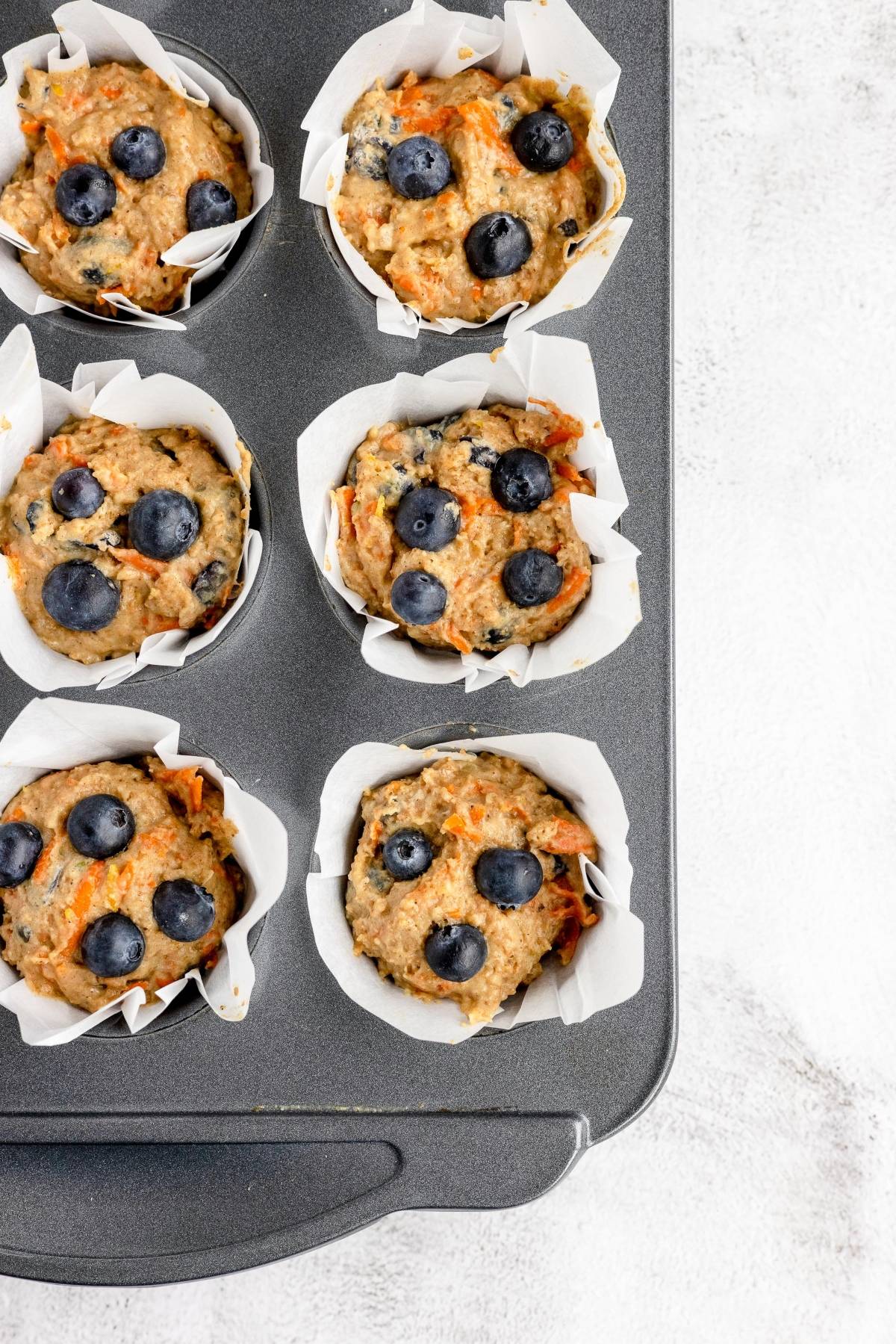 Overhead view of a muffin tin with six carrot blueberry muffins in white paper liners, ready to be baked.