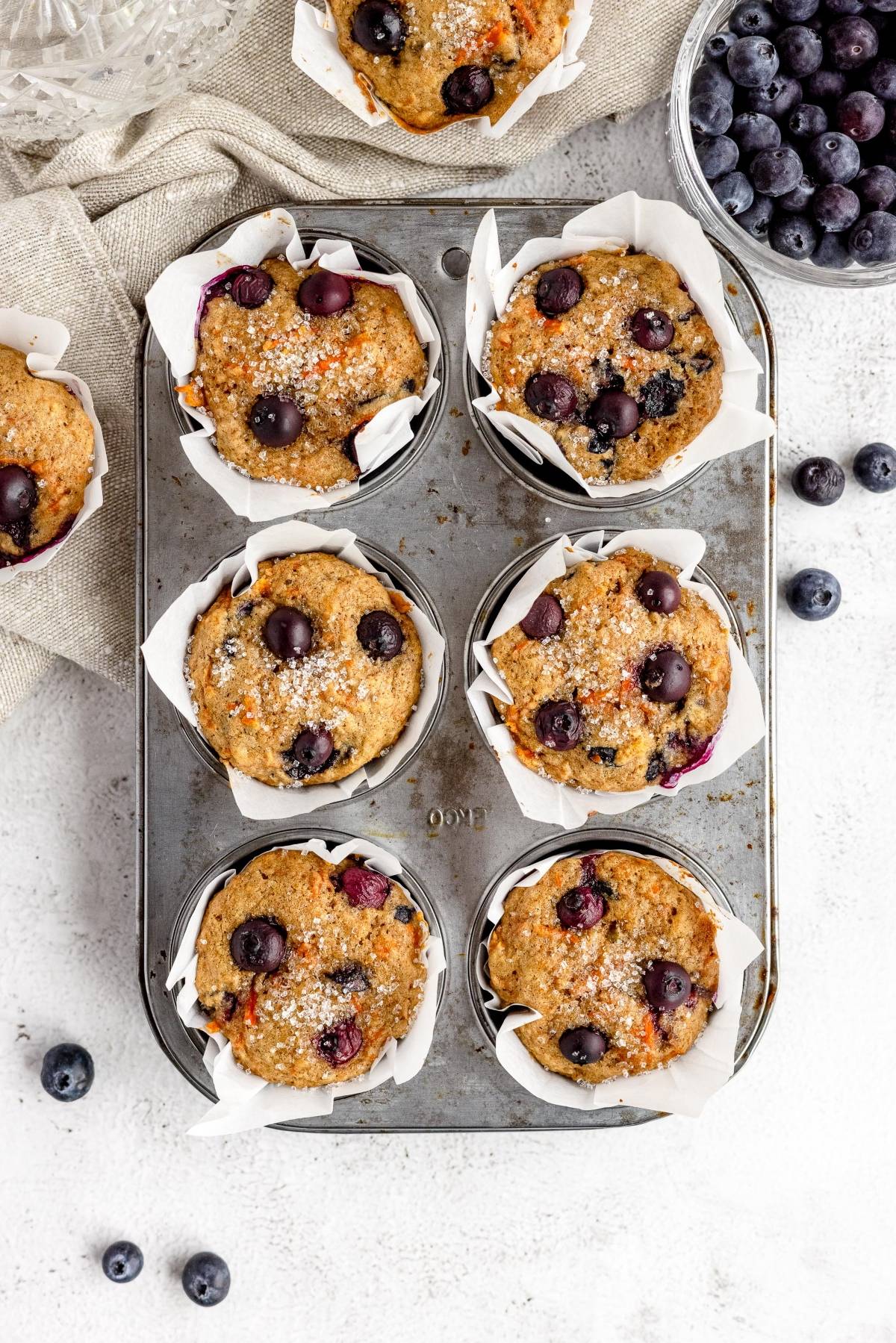 Six carrot blueberry muffins in parchment tulip muffin liners sit in a baking tray, with a bowl of fresh blueberries nearby on a white surface.