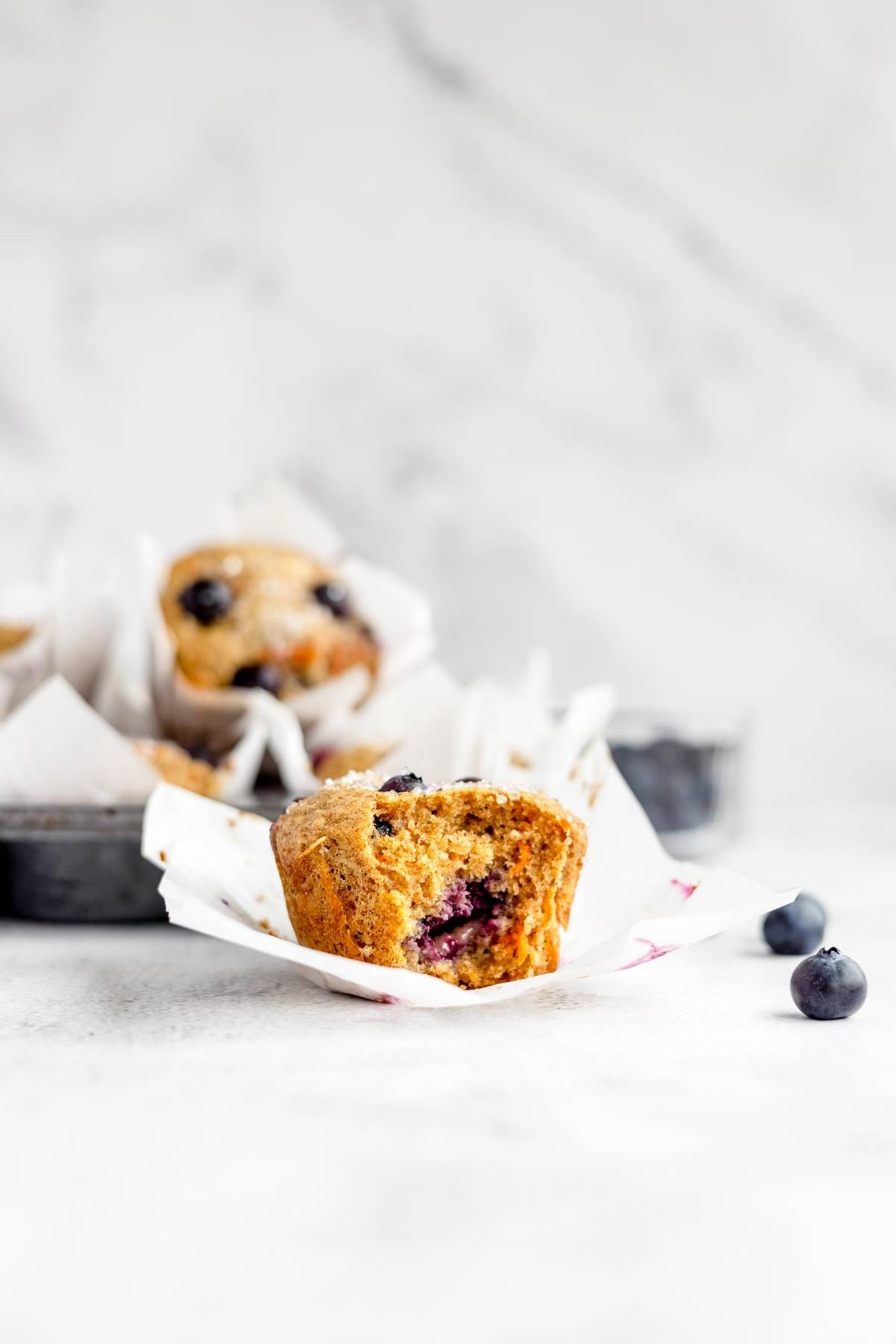 A healthy blueberry muffin with carrots in a paper liner with a bite taken out, with more muffins and blueberries in the background.
