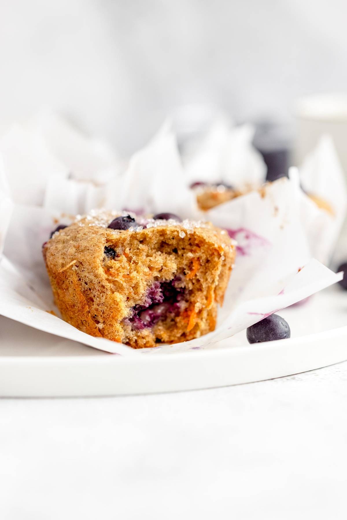 A blueberry muffin with a bite taken out, sitting on white parchment paper on a white plate—perfect alongside carrot blueberry muffins in parchment tulip muffin liners.