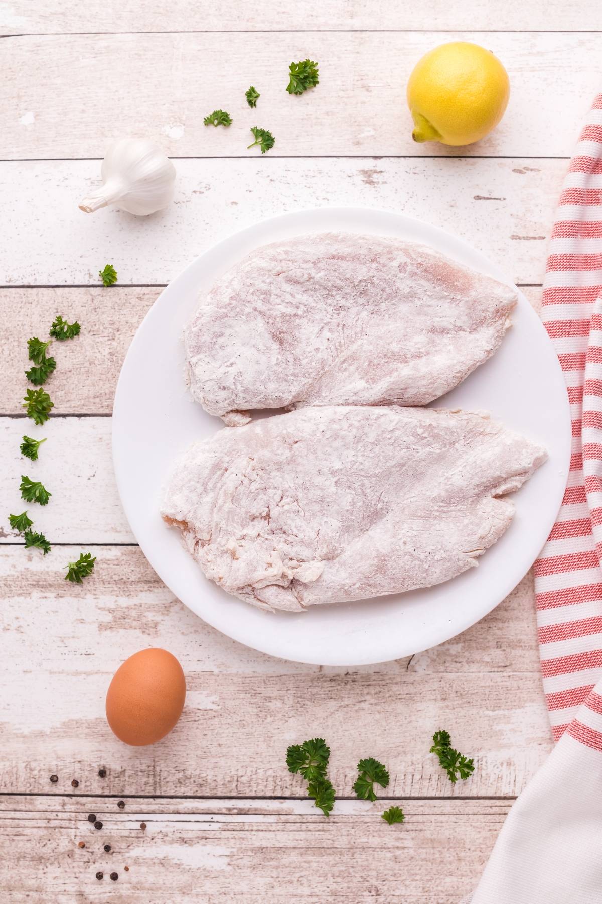 Two raw, breaded chicken breasts on a white plate with an egg, lemon, garlic, and herbs nearby—perfect for preparing Chicken Scallopini with Lemon Sauce, an easy chicken and pasta meal to pair with angel hair pasta.