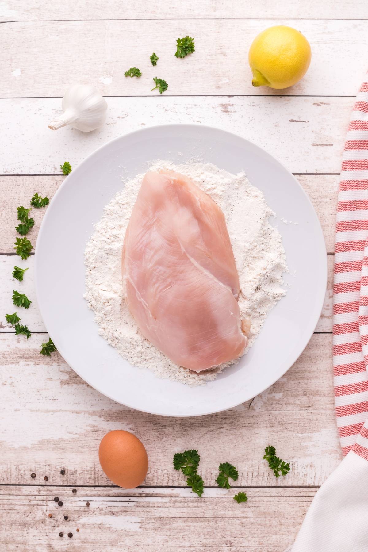 Raw chicken breast on a plate of flour, with egg, lemon, garlic, parsley, and spices on a wooden table—perfect ingredients for an easy Gluten free Chicken piccata or a quick gluten free chicken recipe.