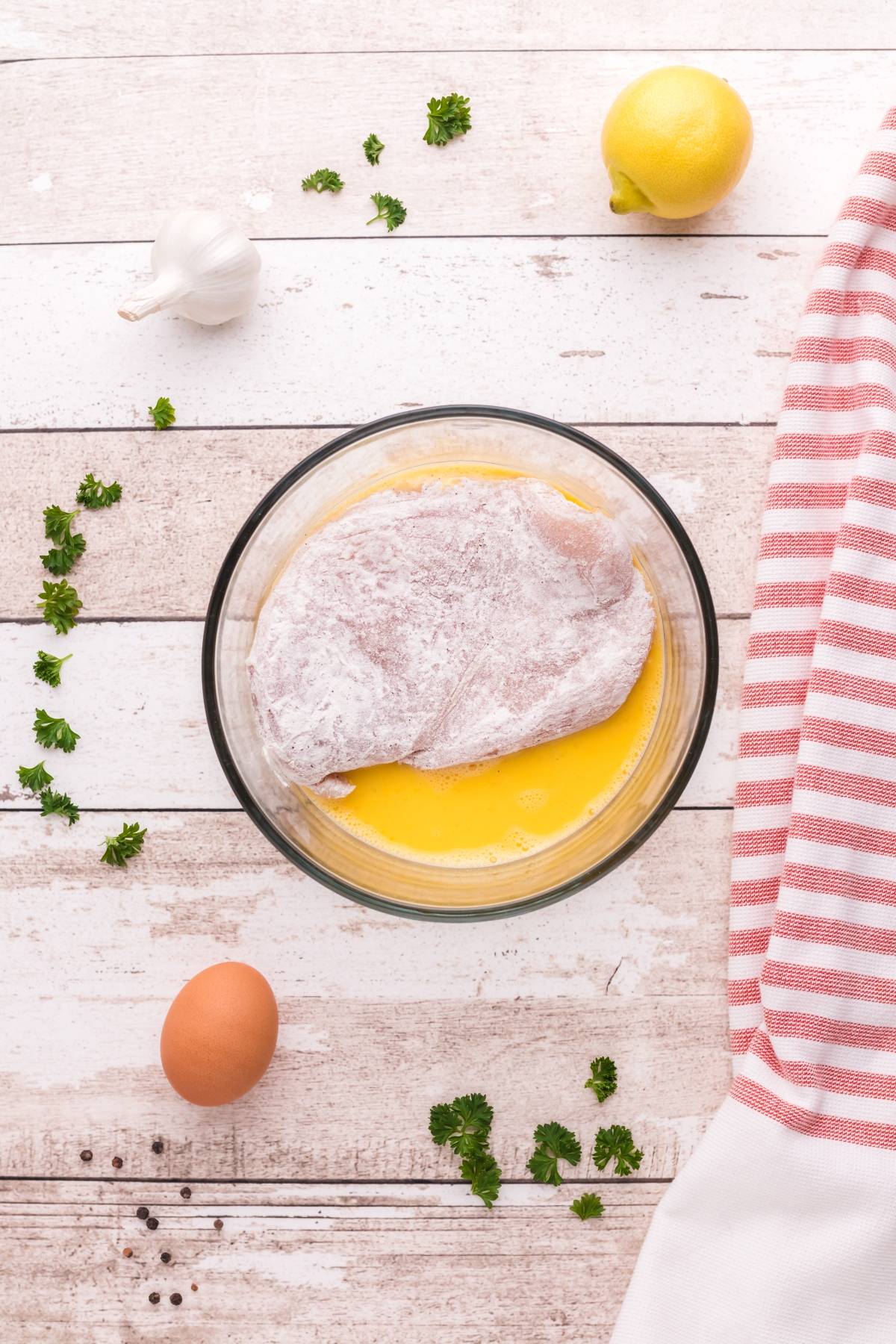 Raw breaded chicken breast in a glass bowl with egg, surrounded by herbs, lemon, garlic, and a striped towel—perfect for preparing Chicken Scallopini with Lemon Sauce or a classic gluten free Chicken Piccata.