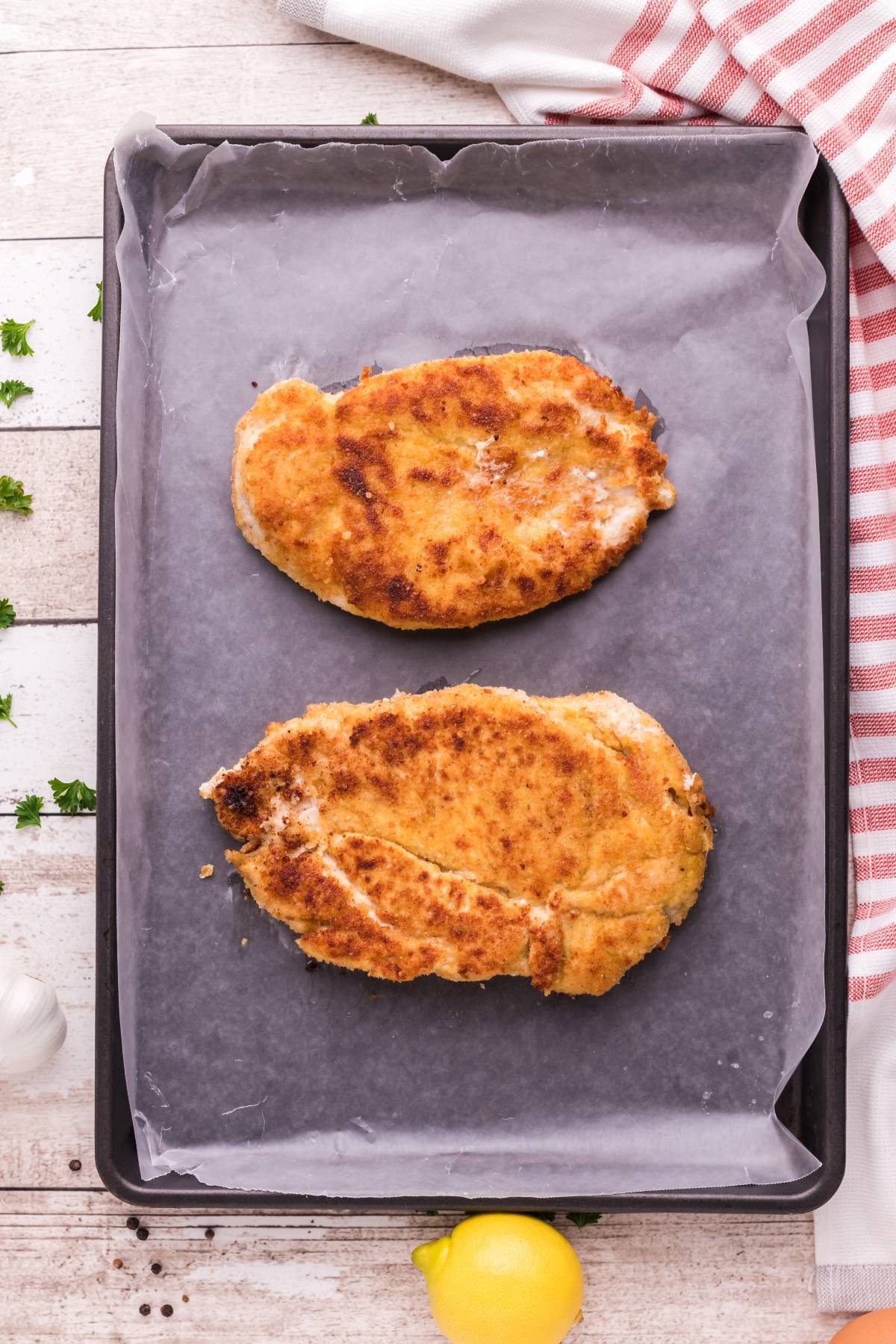 Two breaded chicken cutlets on a parchment-lined baking sheet, perfect for Chicken Scallopini with Lemon Sauce. Serve with angel hair pasta and fresh herbs for an easy chicken and pasta meal bursting with flavor.