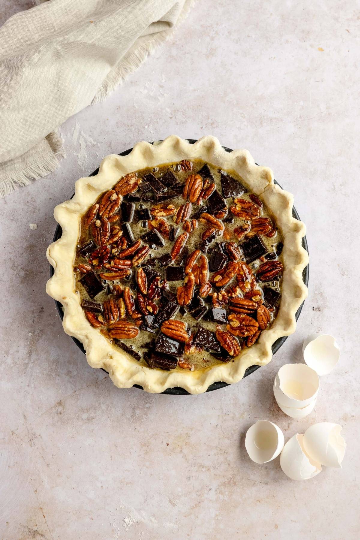 Gooey pecan and chocolate dessert with unbaked chocolate chunks in a pie crust, next to cracked egg shells on a light surface.