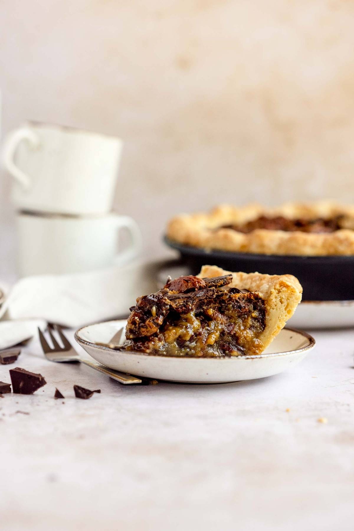 A slice of chocolate pie on a plate with a fork, whole pie and stacked cups in the background—just like the best chocolate pecan pie served at Cracker Barrel, perfect for Thanksgiving or Christmas.