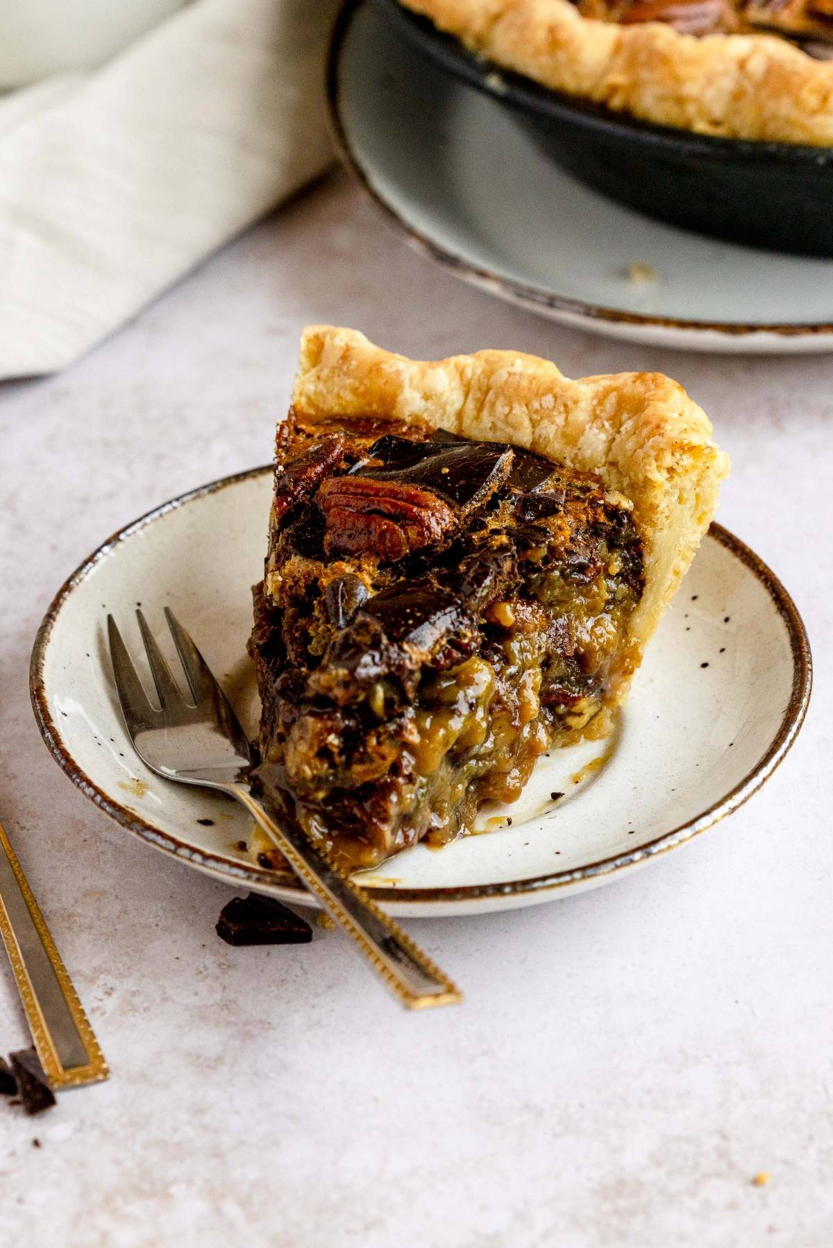 A slice of Chocolate Chunk Pecan Pie on a plate with a fork, showing a gooey filling and flaky crust—perfect as the best chocolate pecan pie to serve for Thanksgiving or Christmas.