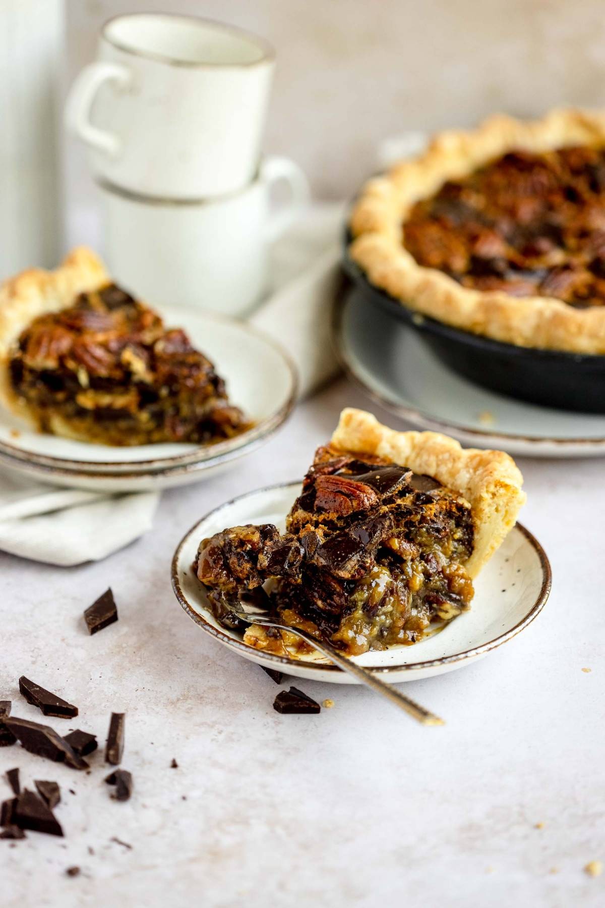 A slice of the best chocolate pecan pie on a plate with a fork, next to the whole pie and some chocolate pieces—a truly gooey pecan and chocolate dessert for any sweet tooth.