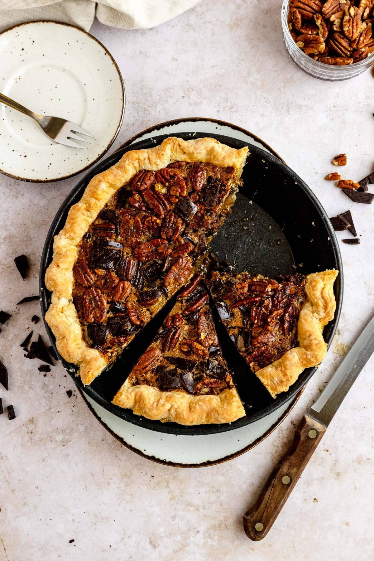 A gooey pecan and chocolate dessert, this pie with two slices missing sits on a plate with a knife, fork, and scattered chocolate and pecans nearby—perfect as the best chocolate pecan pie for Thanksgiving or Christmas gatherings.