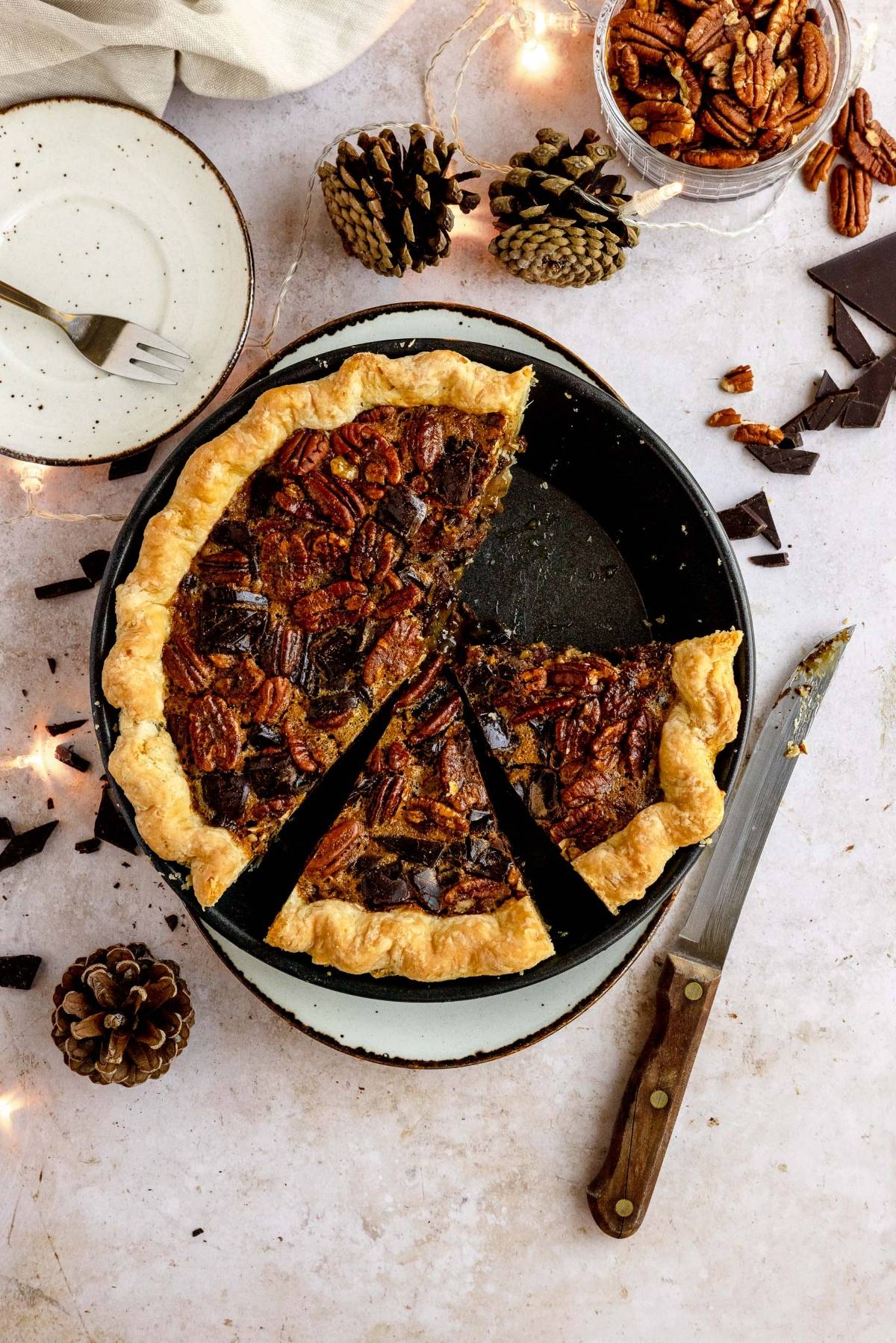 A Chocolate Chunk Pecan Pie with a slice missing, a knife, pinecones, and a plate with a fork on a light surface—perfect for lovers of this gooey pecan and chocolate dessert.