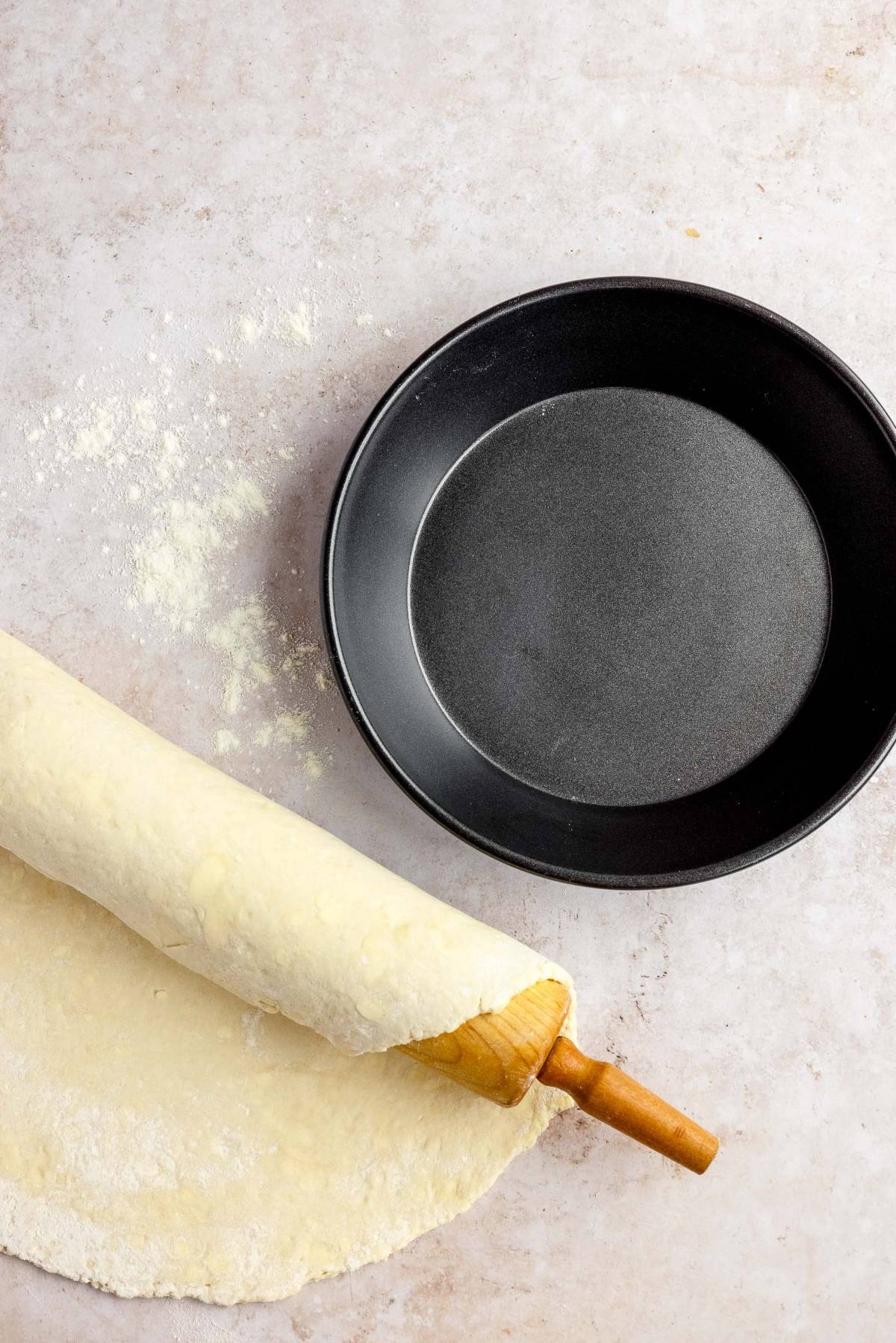 A rolling pin rolls out dough beside an empty, round black pie pan on a floured surface—perfect prep for the Best chocolate pecan pie or any special Pie for Thanksgiving or Christmas.