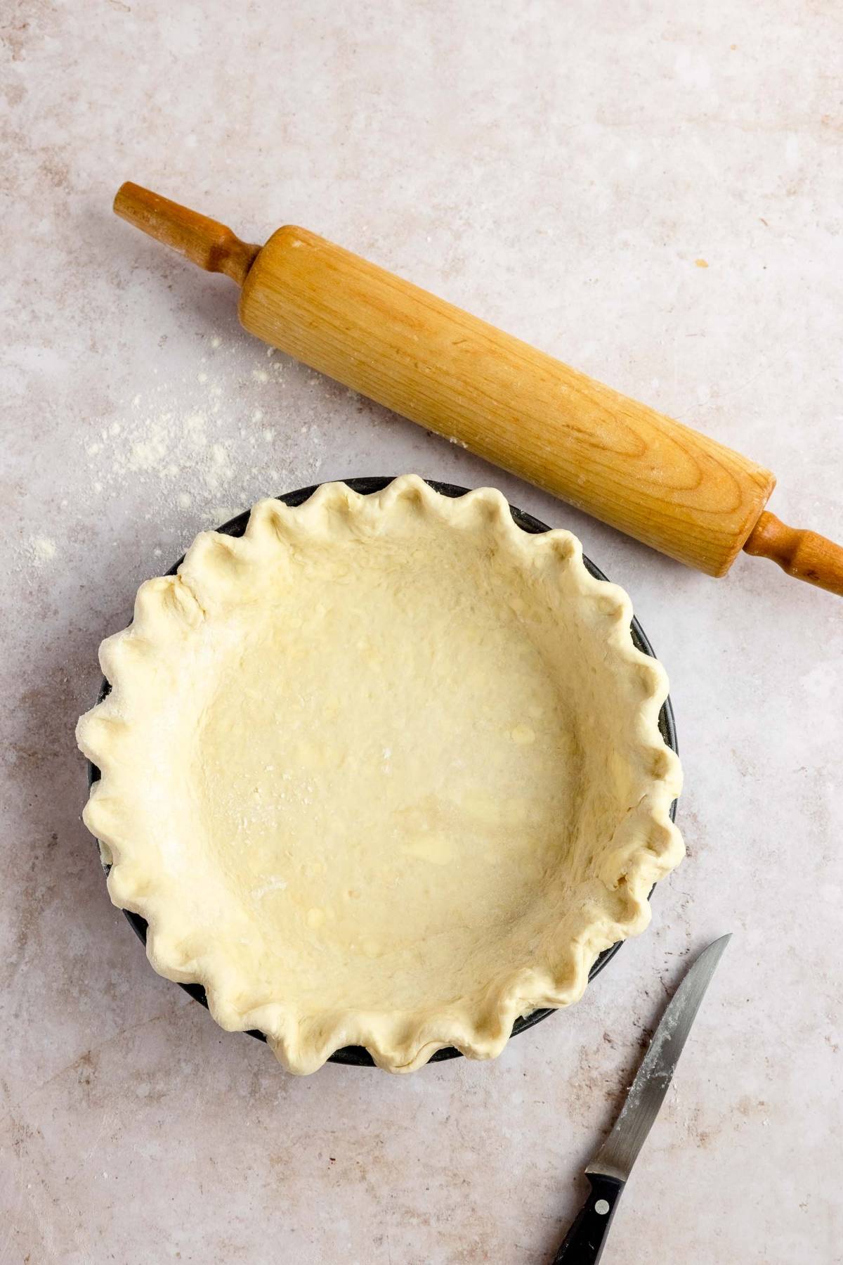 Unbaked pie crust in a pan with fluted edges, rolling pin, and paring knife on a light surface—perfect for preparing the best chocolate pecan pie or any gooey pecan and chocolate dessert.