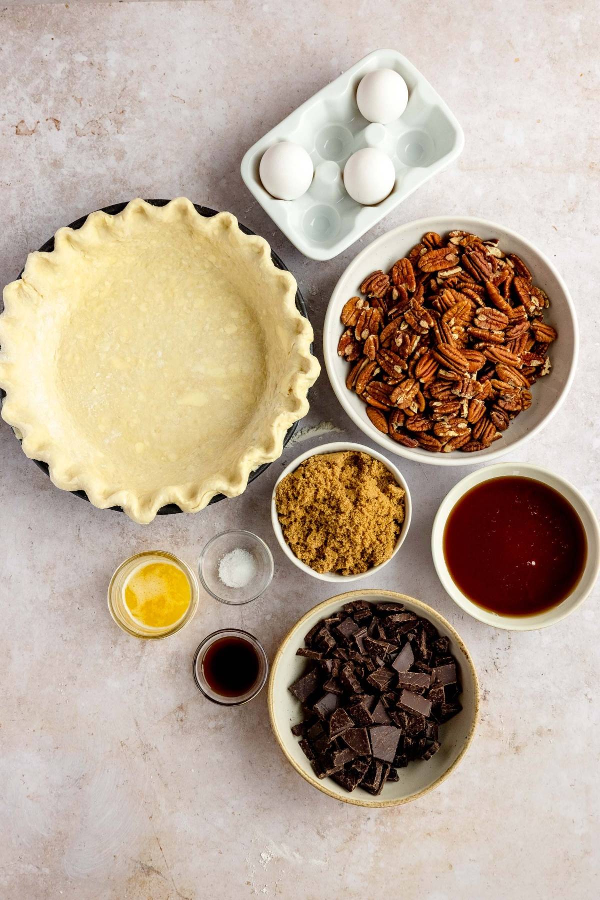 Unbaked pie crust, eggs, pecans, brown sugar, chocolate chunks, syrup, butter, vanilla, and salt on the counter—everything you need for a gooey pecan and chocolate dessert like Chocolate Chunk Pecan Pie for Thanksgiving or Christmas.