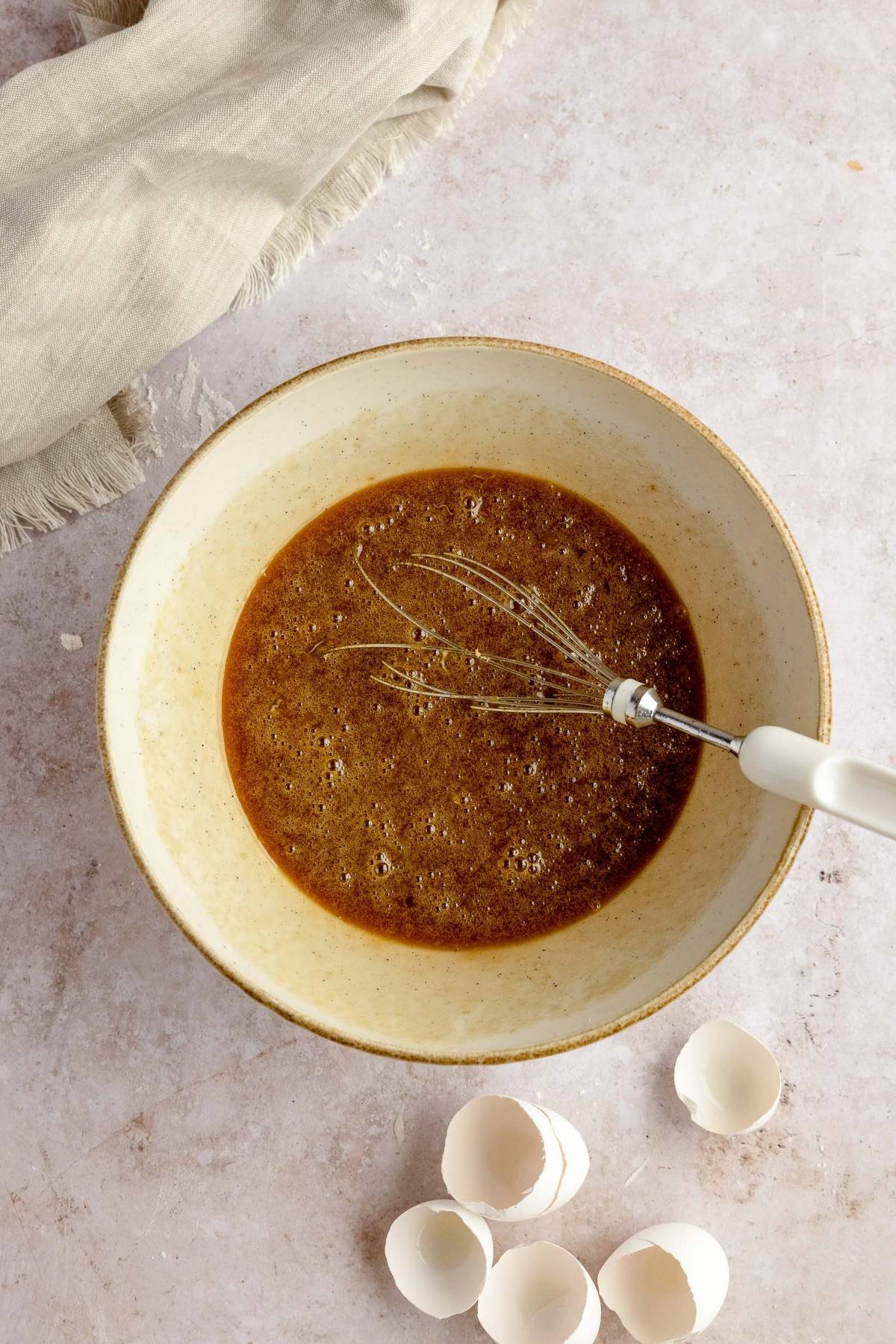 A whisk in a bowl of brown batter with empty eggshells and a beige cloth nearby on a light surface, ready to become a gooey pecan and chocolate dessert like Cracker Barrel pecan pie.