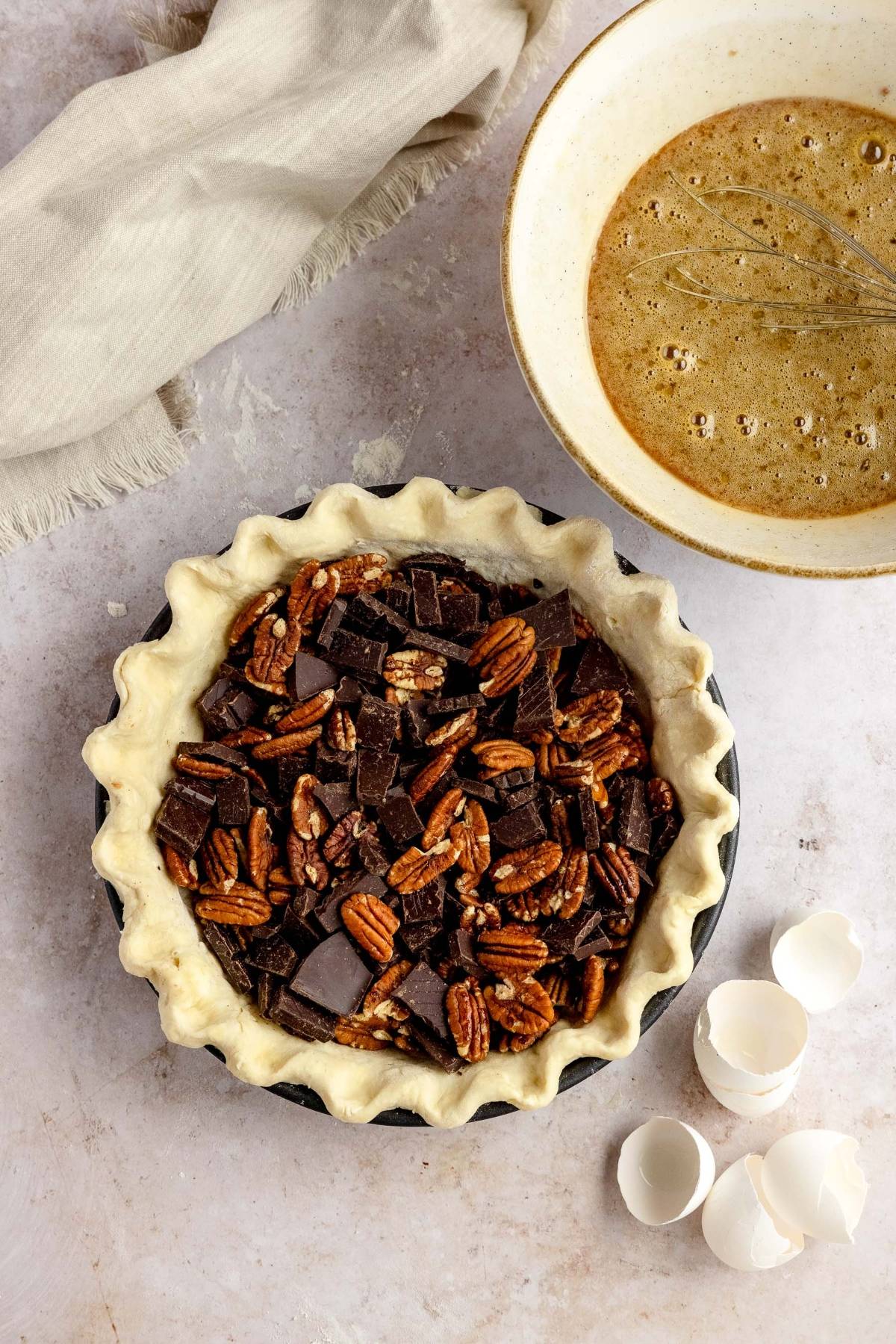 Unbaked Chocolate Chunk Pecan Pie—just like Cracker Barrel pecan pie—with chunks in a flaky crust, sits beside a bowl of filling and broken eggshells.