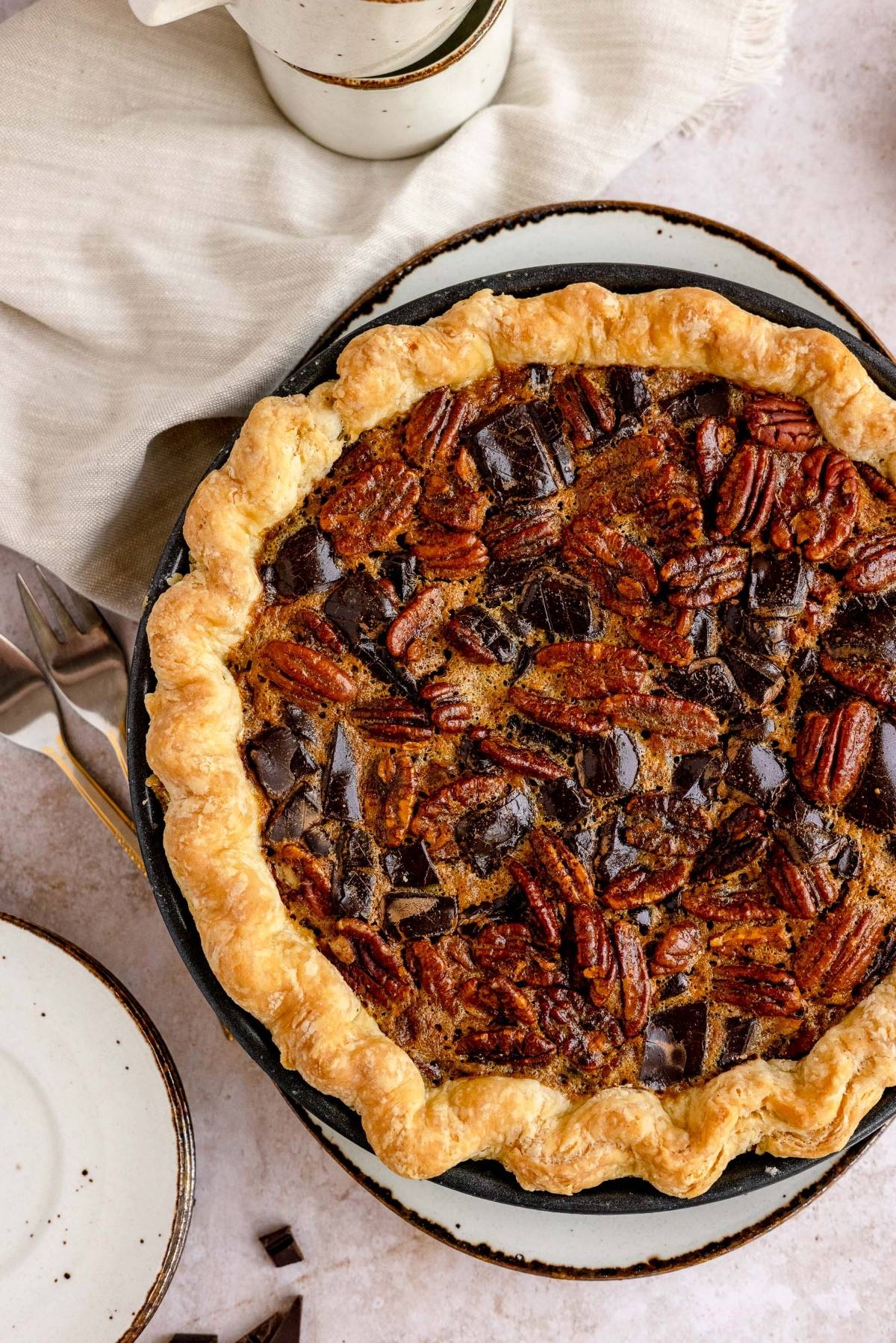 A pecan pie with chocolate chunks in a flaky crust on a plate, just like the best chocolate pecan pie you’d find at Cracker Barrel—perfect as a pie for Thanksgiving or Christmas, served next to a fork and napkin.