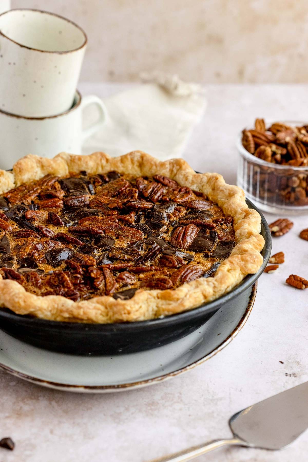 A baked Chocolate Chunk Pecan Pie sits on a plate beside a bowl of pecans and stacked cups on a light background.