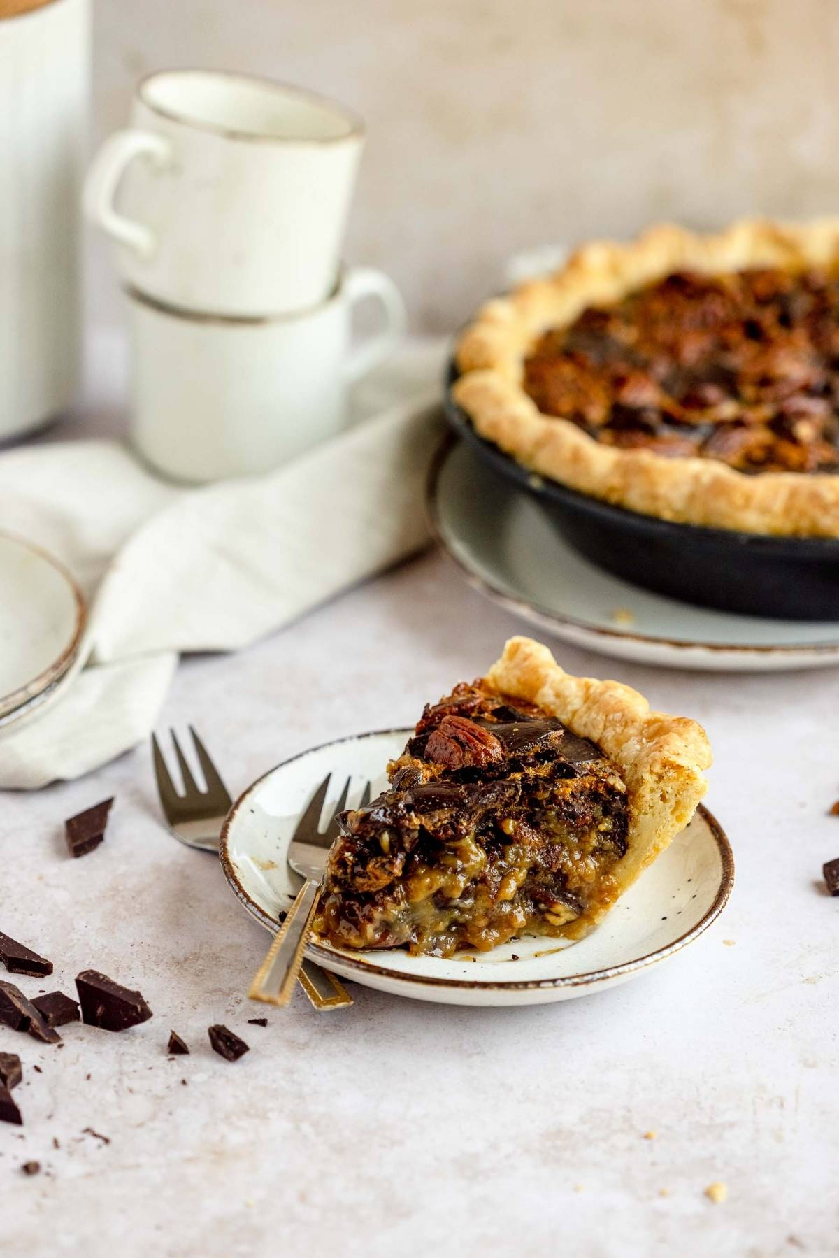 A slice of Chocolate Chunk Pecan Pie on a plate with a fork, next to the whole pie and stacked cups in the background—this gooey pecan and chocolate dessert is sure to be your new favorite!.