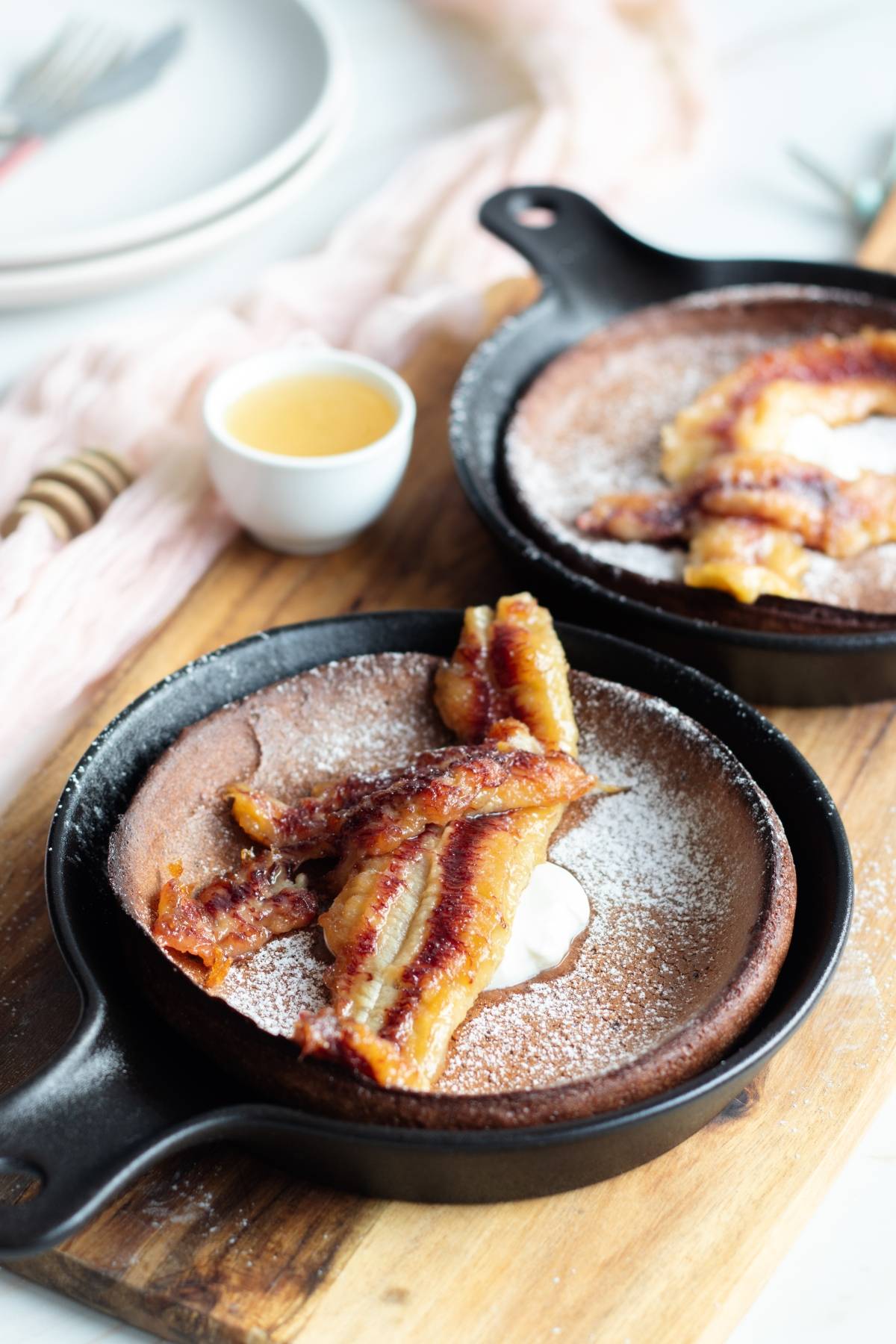Two mini skillets with chocolate dutch baby pancakes, topped with caramelized bananas and powdered sugar, served on a wooden board.