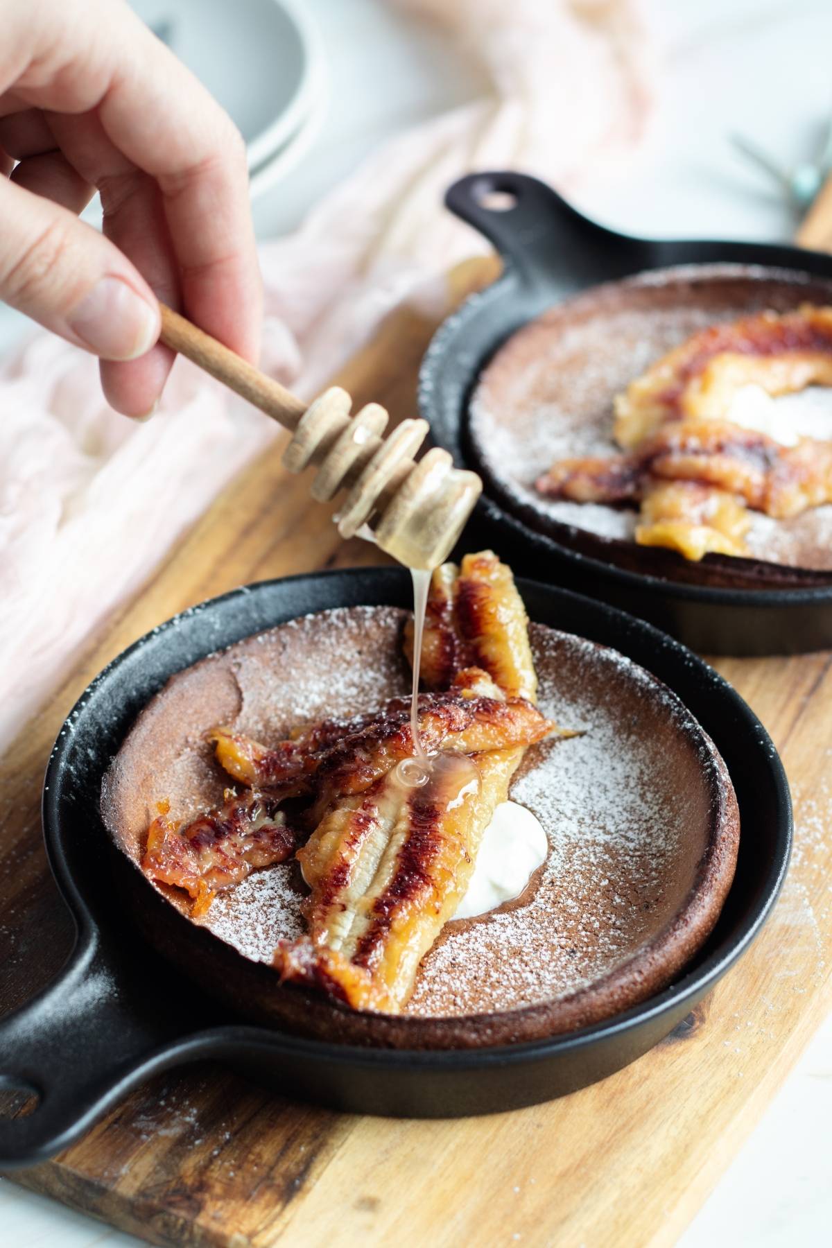 A hand drizzles honey over baked bananas and cream atop a Chocolate Dutch Baby in a small cast iron skillet dusted with powdered sugar.