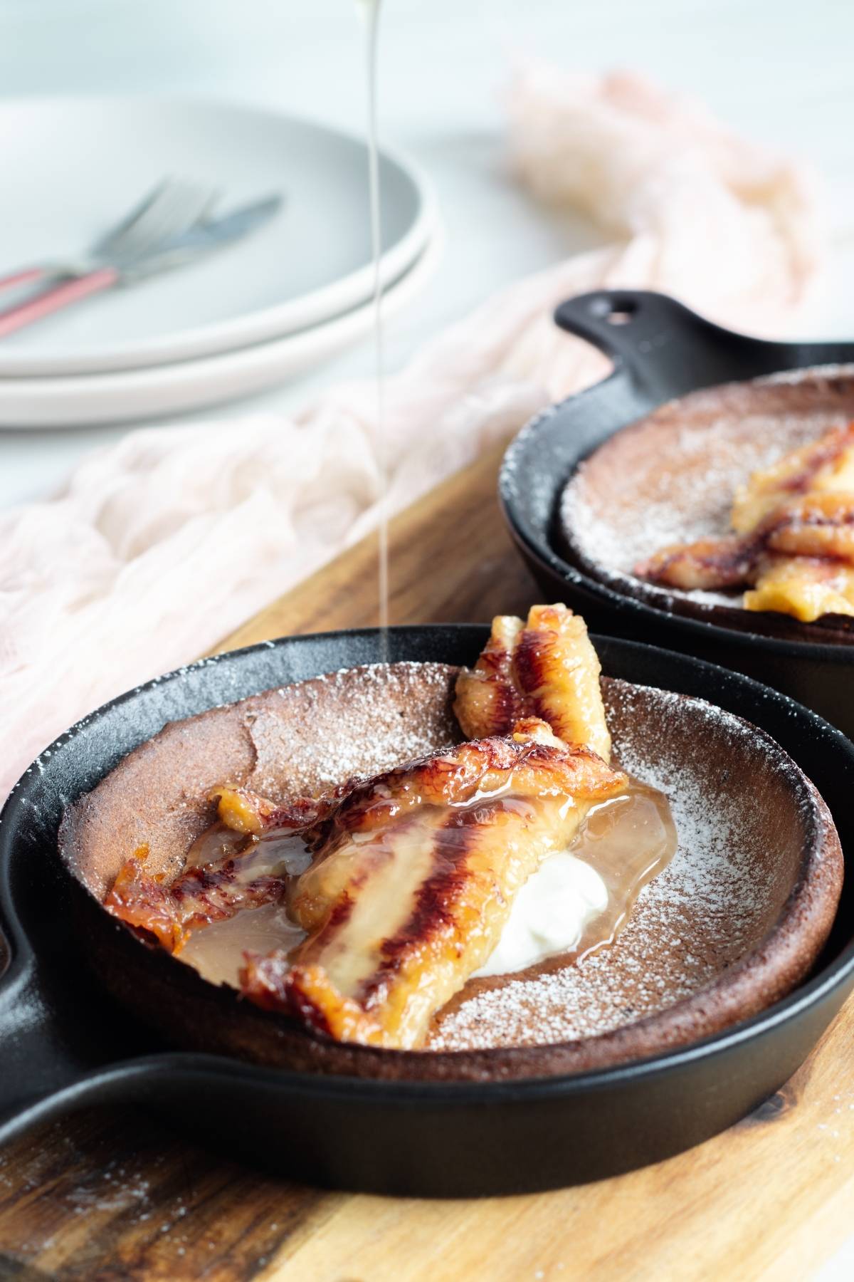 Caramelized banana in a skillet with powdered sugar and sauce being poured on top, perfect for topping a gluten free dutch baby. Plates and forks in the background.