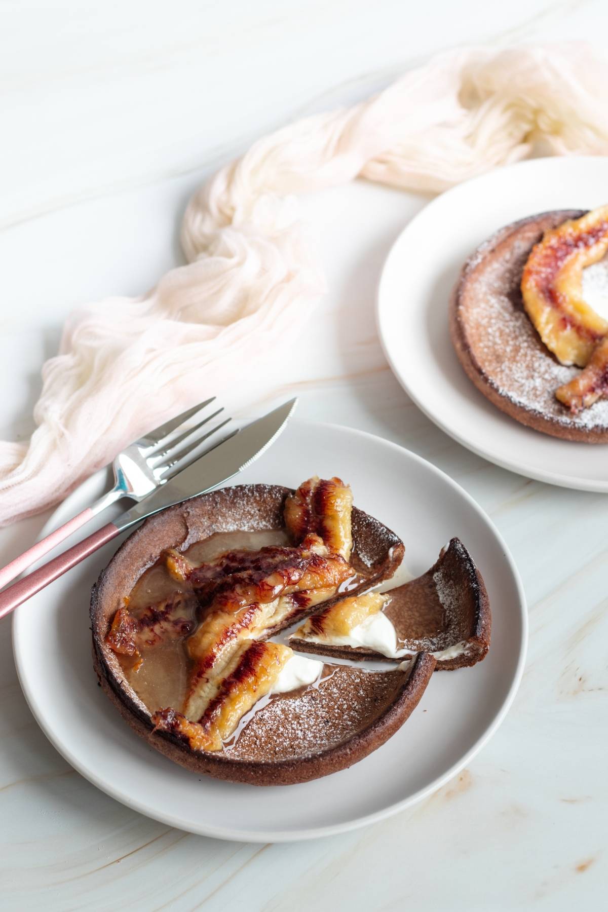 Gluten free Dutch baby pancake with caramelized bananas and cream on a white plate, with a fork and knife nearby.