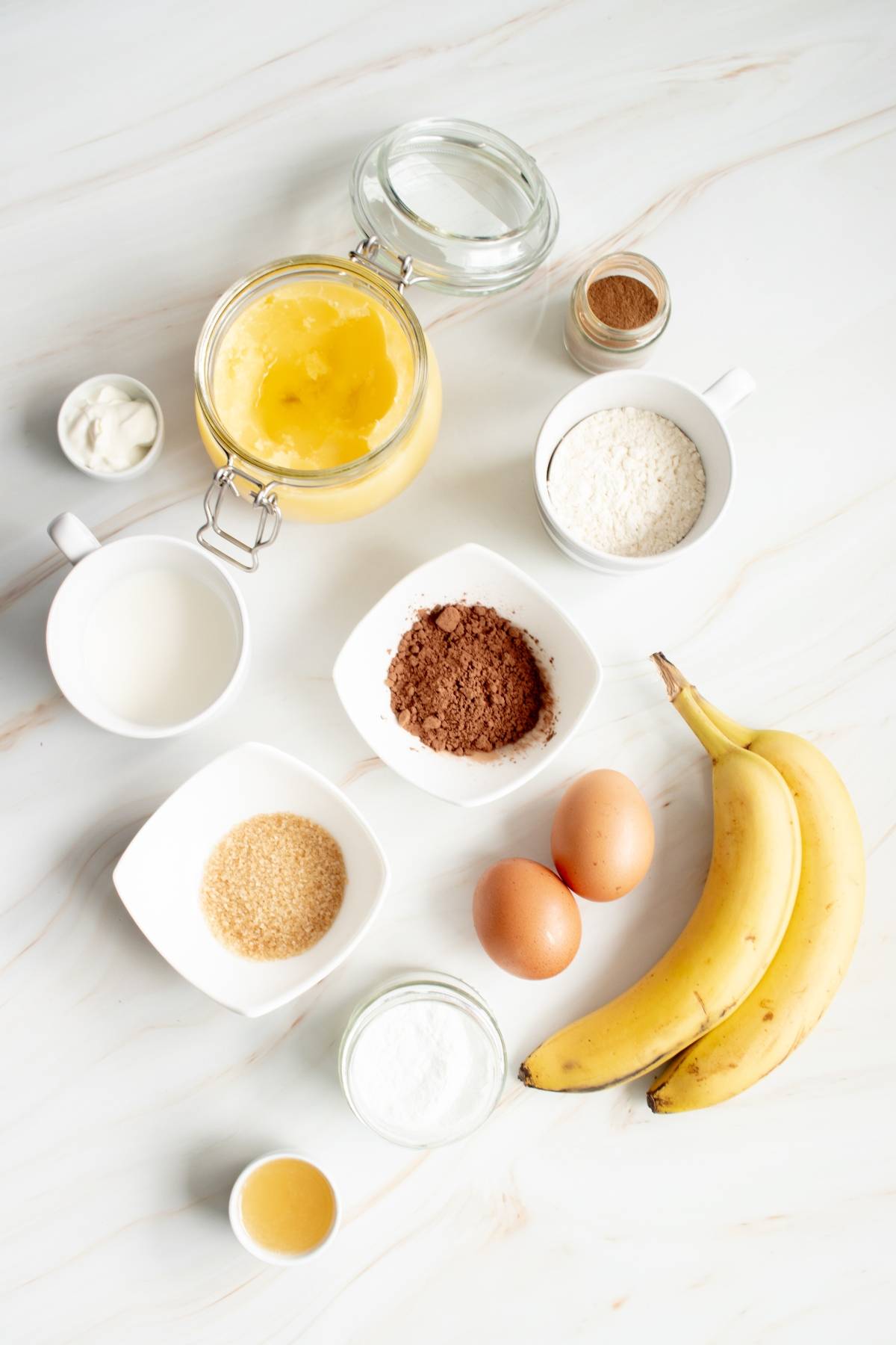 Baking ingredients on a white surface: bananas, eggs, flour, cocoa, sugar, milk, butter, and spices—everything you need for a delicious Chocolate Dutch Baby or your favorite Chocolate German pancake recipe.