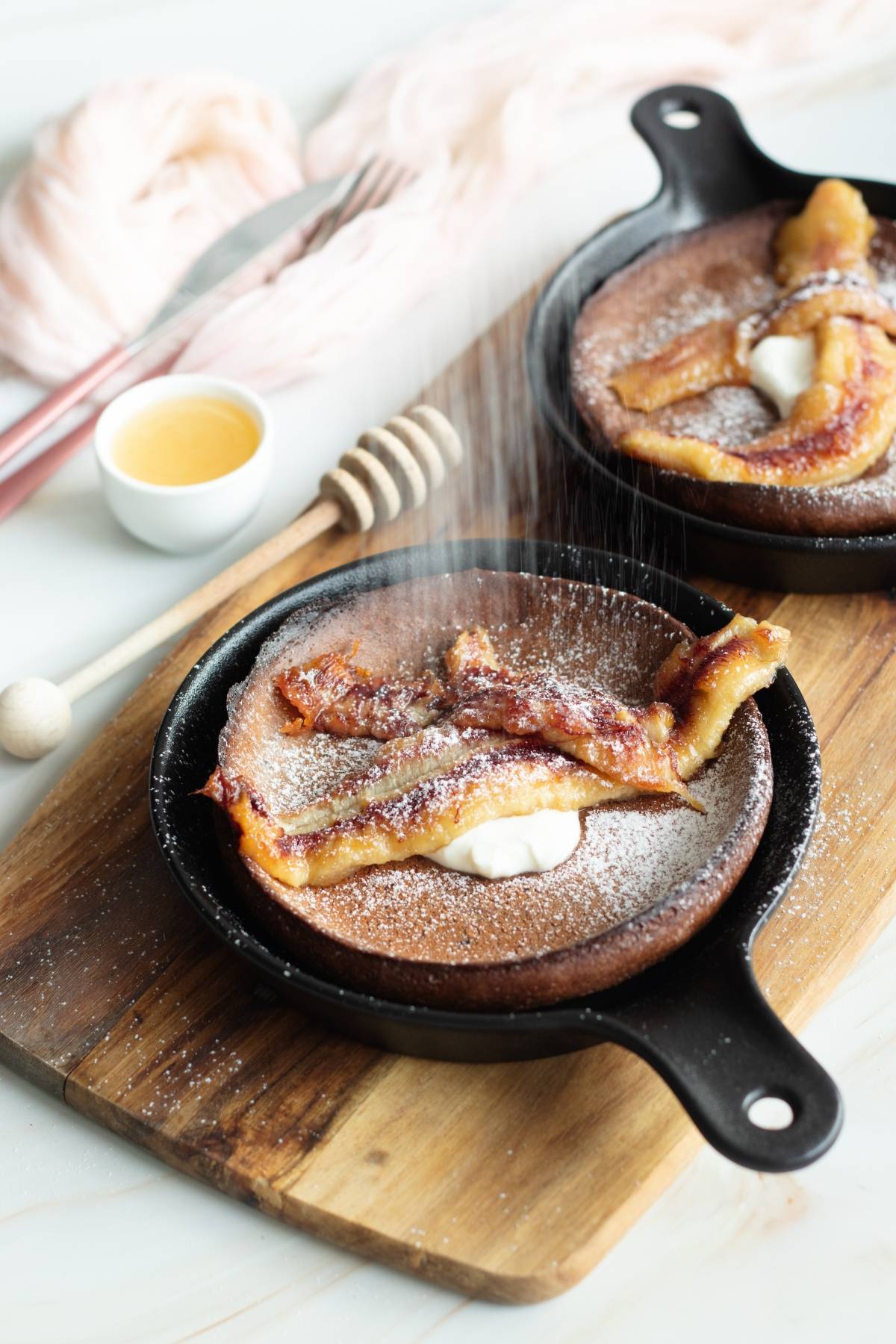 Mini gluten free Dutch baby pancake topped with caramelized bananas, powdered sugar, and a dollop of cream on a wooden board.