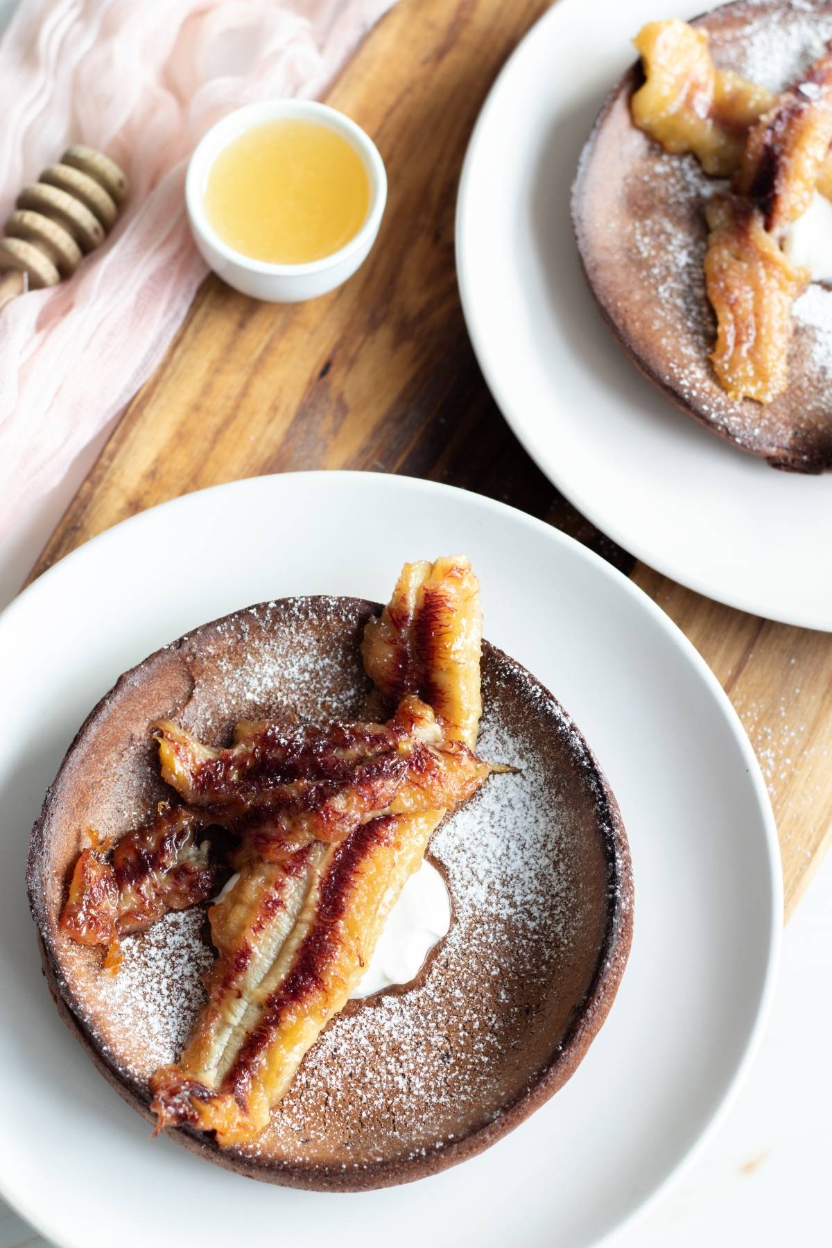 Chocolate German pancake recipe: Chocolate pancakes topped with caramelized bananas and powdered sugar on white plates, with honey in a small bowl. Perfect for those seeking a delicious twist on the classic Chocolate Dutch Baby.