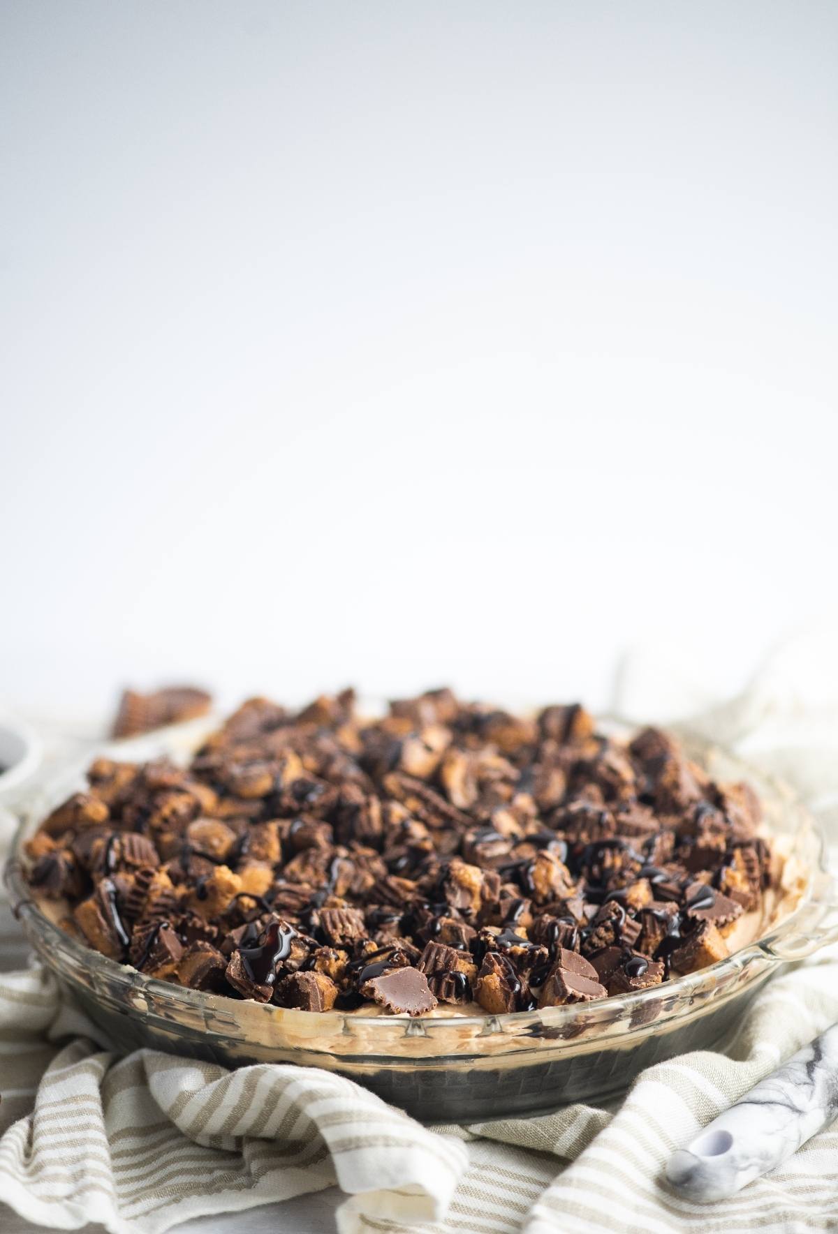 A chocolate peanut butter pie topped with chopped peanut butter cups and chocolate drizzle rests on a striped cloth.