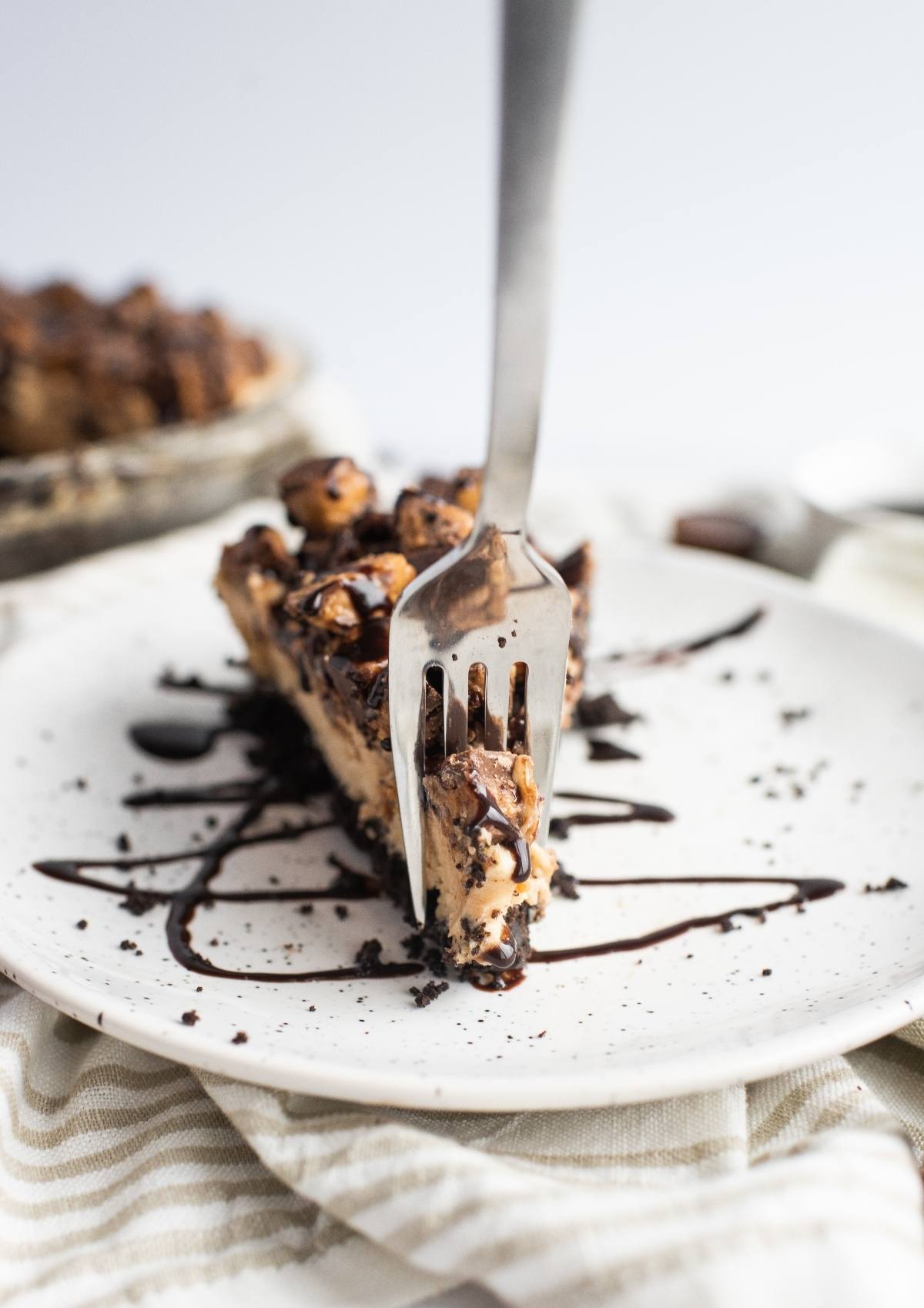 A fork cuts into a slice of chocolate-drizzled chocolate peanut butter pie on a white plate.