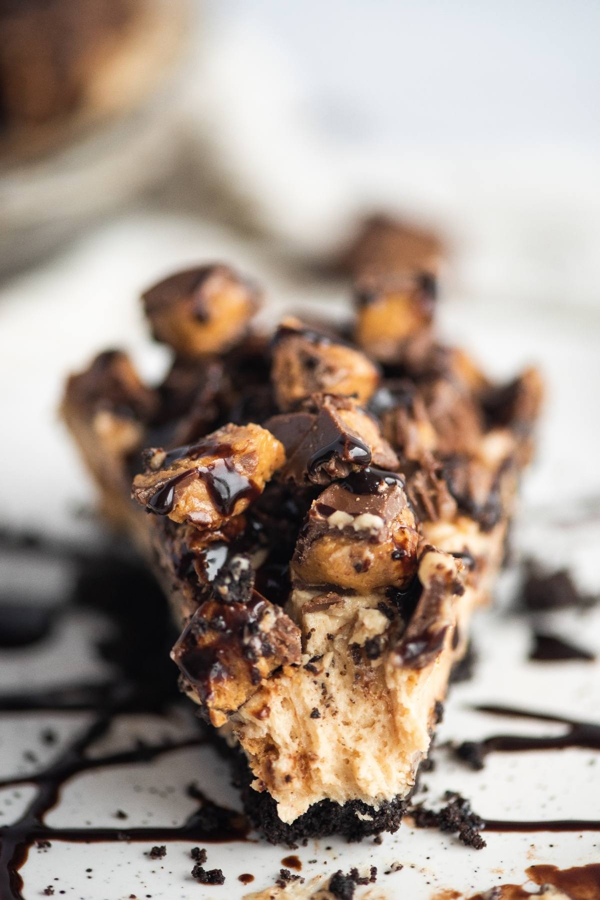 A slice of peanut butter cream pie with a chocolate graham crust, topped with chopped peanut butter cups and rich chocolate drizzle.
