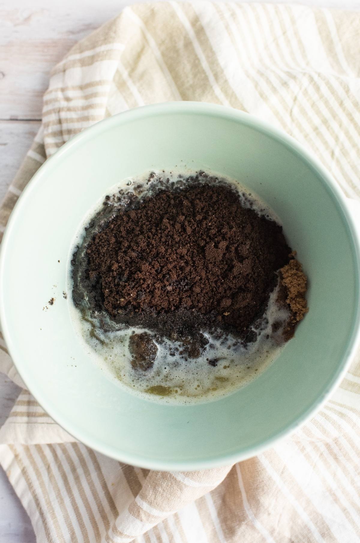 A mixing bowl with melted butter, brown sugar, and crushed chocolate cookies sits on a striped cloth—just the beginning steps for a decadent chocolate graham crust in a classic chocolate peanut butter pie.